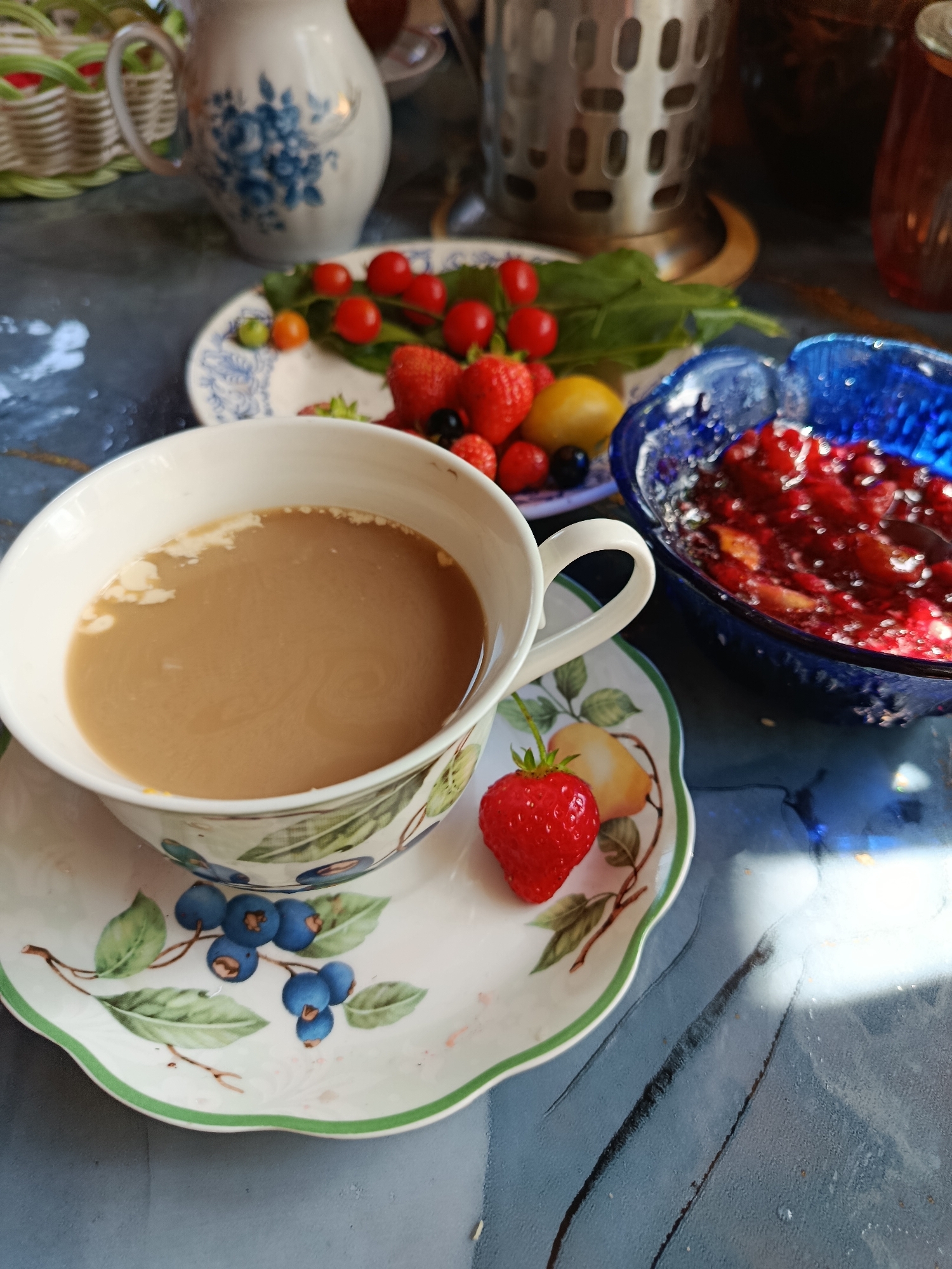 The image depicts a close-up, eye-level view of a table setting featuring a cup of coffee and various fruits. A white porcelain cup and saucer, adorned with a painted pattern of blueberries and leaves, sits prominently in the foreground. The cup contains a light brown beverage, presumably coffee with milk, showing a swirling pattern on its surface. A single red strawberry is placed on the saucer.
Behind the cup, a small white plate holds a cluster of red cherry tomatoes, a few small green berries, and several ripe strawberries. A bright yellow lemon or small apple is also visible on this plate, along with some green leaves.
To the right of the cup, a vibrant blue glass bowl is filled with a red fruit compote or jam, appearing to contain pieces of fruit. The background is softly blurred, but suggests a rustic or outdoor setting with a woven basket and a metal container, possibly a pitcher or a utensil holder. Sunlight creates a dappled effect on the textured surface of the table, which appears to be a dark blue or grey material.
The scene is positioned on a table, likely outdoors or near a window, given the quality of light. The overall impression is one of a relaxed breakfast or snack. There are no people visible in the image. The location is not specified as being in the city of Верея, Russia; this detail is not supported by visual evidence in the image itself.