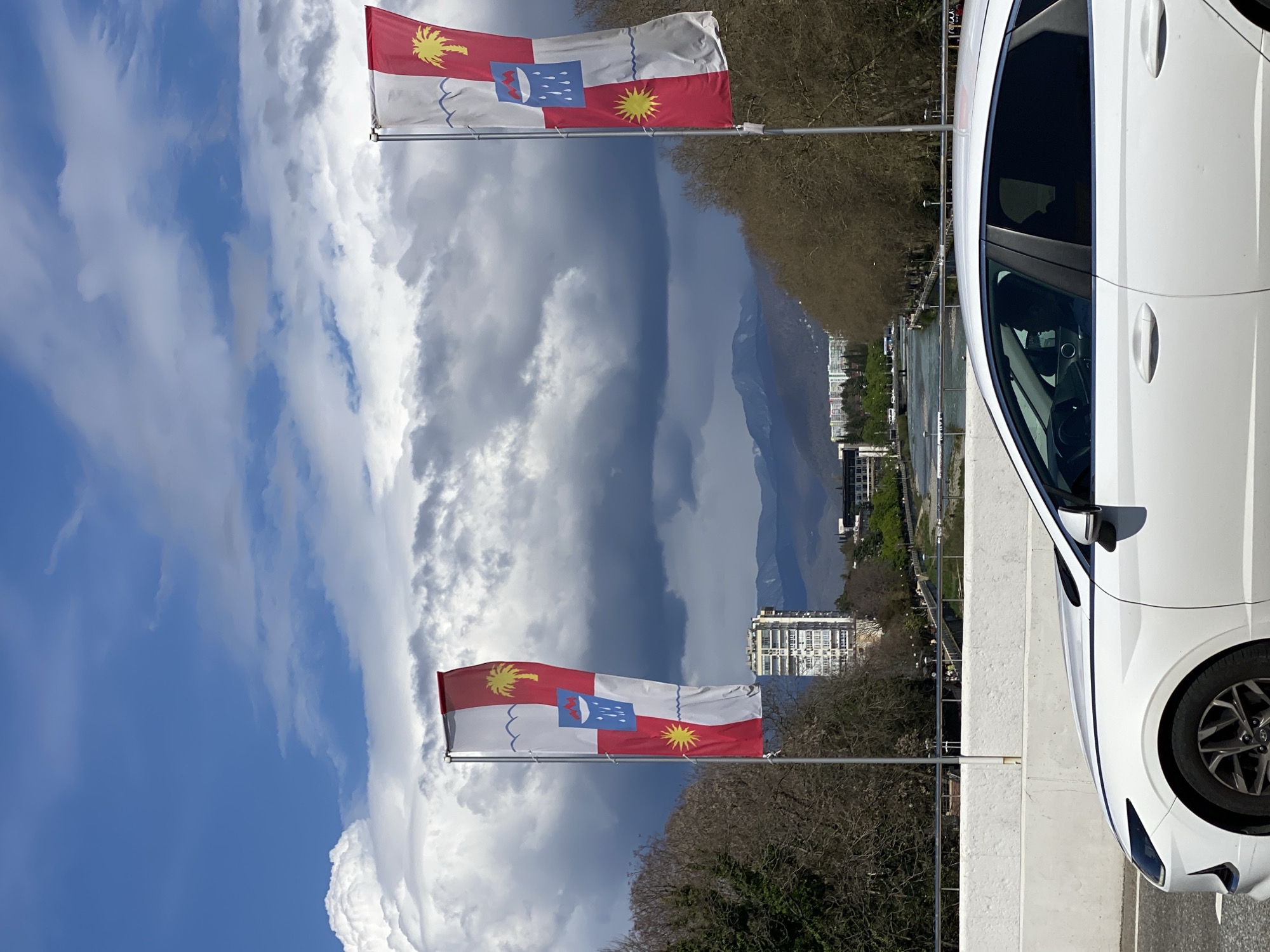 The image presents an outdoor scene featuring the flag of Abkhazia waving in front of a cloudy sky and a white car partially visible on the right. In the background, there is a body of water, potentially a river or lake, with buildings and trees lining its banks. Mountains are visible in the distance. There's no information that allows me to verify that this is Sochi, Russia. It depicts flags, vehicles, and landscapes without any specific details or actions.