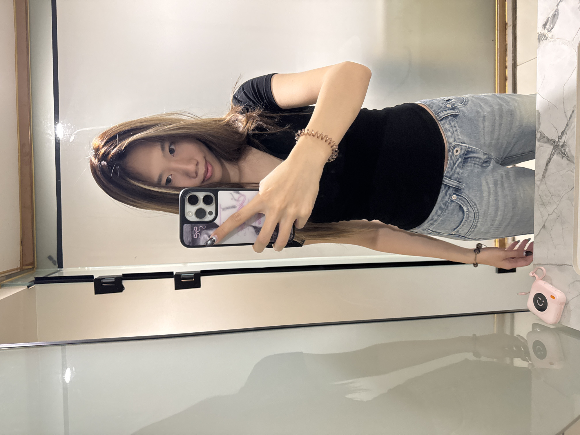 A young woman is taking a selfie in a bathroom. She is holding a smartphone with a patterned case and her face is partially visible in the reflection. She is wearing a black shirt, light blue jeans, and a spiral hair tie on her wrist. A white sink counter and a pink mini printer are visible in the foreground. The reflection indicates a modern, clean bathroom setting. While specific location details for the Yuzhong District, China are not readily apparent from the image, the bathroom appears to be located in a commercial or residential building.