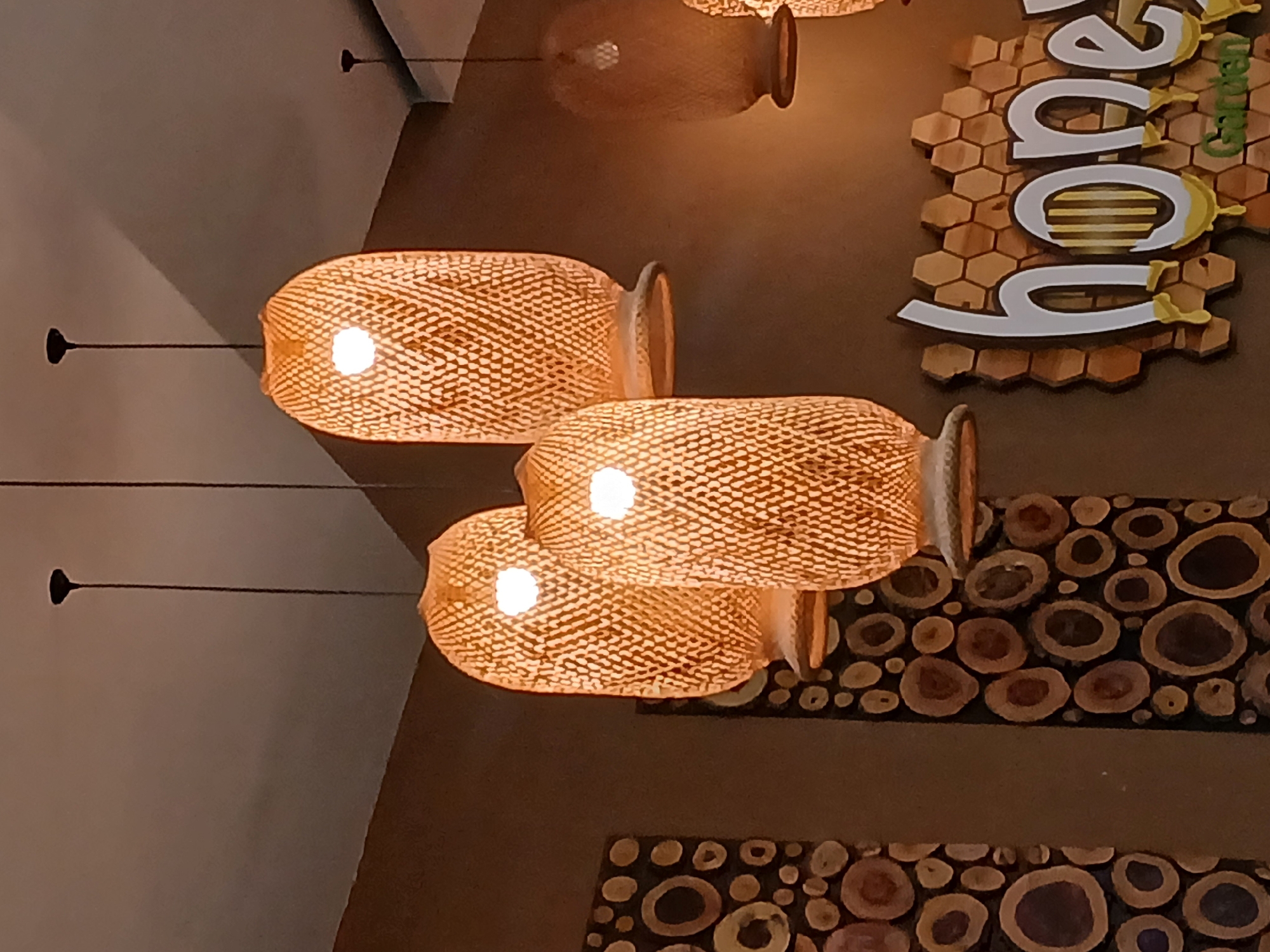 The image displays the interior of the Honey Garden Café, situated within the Los Olivos Shopping Center (C.C. Los Olivos) in Puerto Ordaz, Ciudad Guayana, Venezuela. Three primary woven rattan pendant lamps with internal illumination hang from a light-colored ceiling, casting a warm orange glow against a dark brown wall. A fourth similar lamp is partially visible on the far right. Two vertical decorative panels on the left wall are adorned with various-sized circular wood slices, exposing their natural concentric ring patterns. On the right wall, a multi-layered sign is mounted, featuring a honeycomb pattern as its base. Overlaid in white stylized lettering are the characters "hone" with yellow drip accents, with a partial view of a 'y'. Below this, smaller green text reads "Garden." No individuals or active interactions are present in the scene.