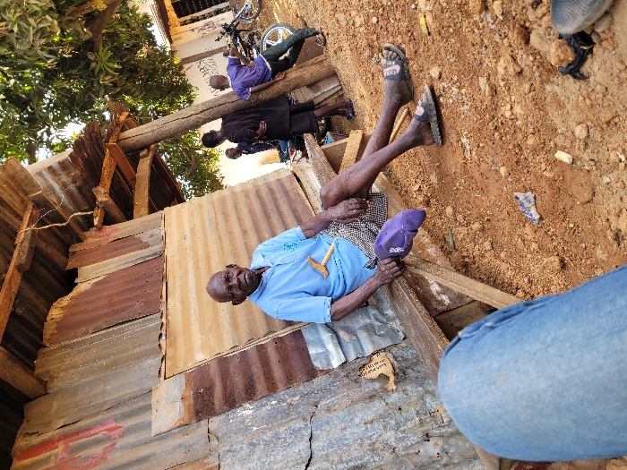 The scene is set outdoors in Ube, Nigeria. An elderly, bald man with dark skin sits on a simple wooden bench, leaning against a wall constructed from various corrugated metal sheets, some rusted. He wears a light blue polo shirt, patterned shorts, and dark sandals. His right hand holds a light-colored wooden object, and a purple cap rests on the bench near his left leg. His legs are extended, with his feet resting on the reddish-brown, dusty ground, which is sparsely littered with small debris, including a white and blue wrapper.
Behind and to the right of the seated man, a wooden pole supports a corrugated metal overhang. Three other individuals are visible in the background: two men in dark shirts stand, partially obscured, and a third man in a purple shirt and light trousers leans against the wooden pole, facing away from the camera. Further in the background, a light-colored building with dark windows or doorways is visible, and a motorcycle is parked nearby. A large tree with green leaves occupies the upper right portion of the frame, casting shadows. The image is captured from a low vantage point, with the photographer's blue-jeaned legs visible in the bottom left foreground.