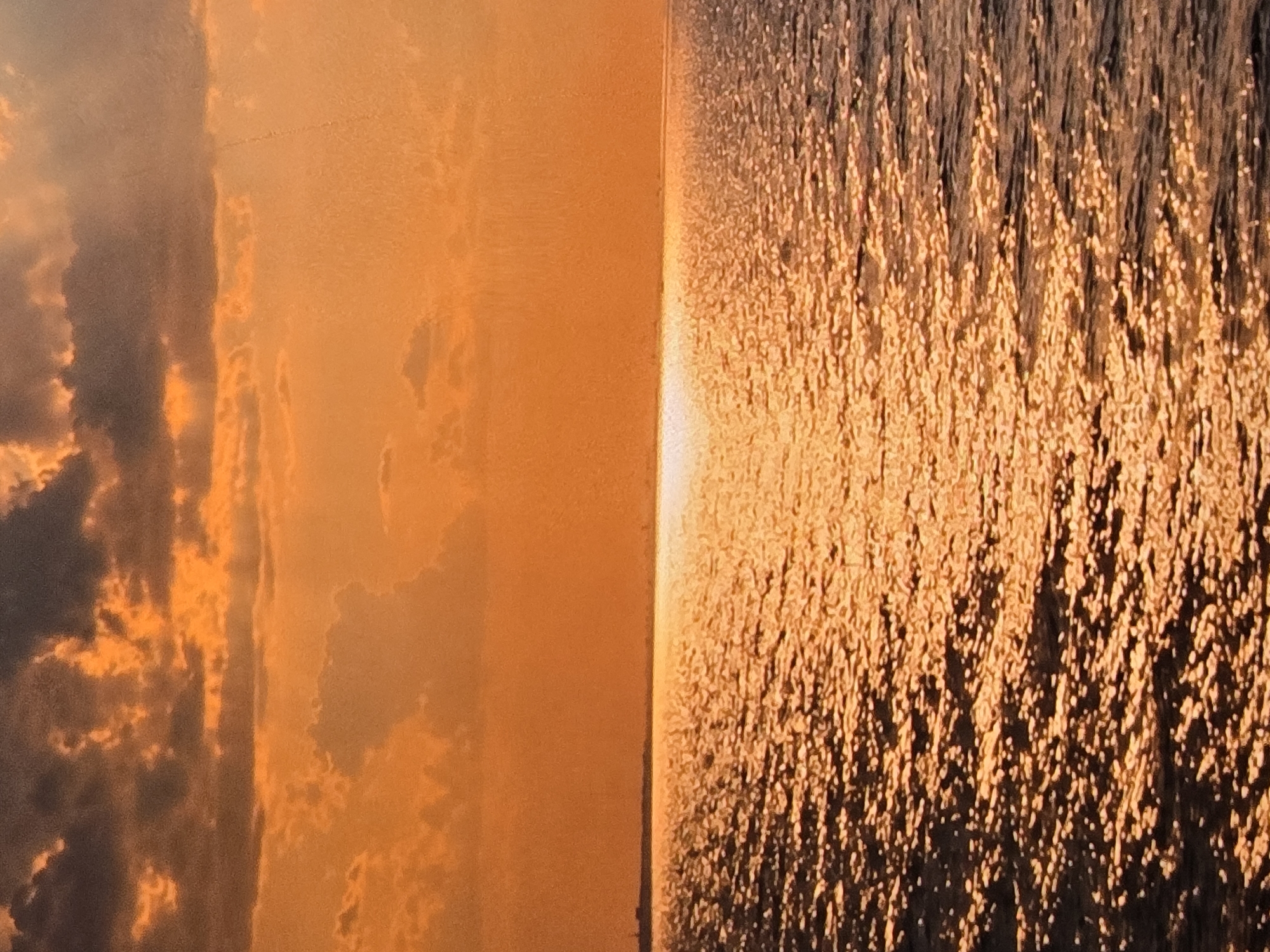 This is a vertical, eye-level, medium shot photograph taken at sunset over a body of water. The sky is filled with dramatic clouds, tinged with oranges and grays, with the sun's rays filtering through. The sun itself is a bright horizontal streak of light on the horizon, reflecting off the rippling water. The water appears choppy, with individual waves catching the light. In the far distance, a faint, dark line suggests land or a distant city skyline. This image does not depict any location in the city of Vladimir, Russia.