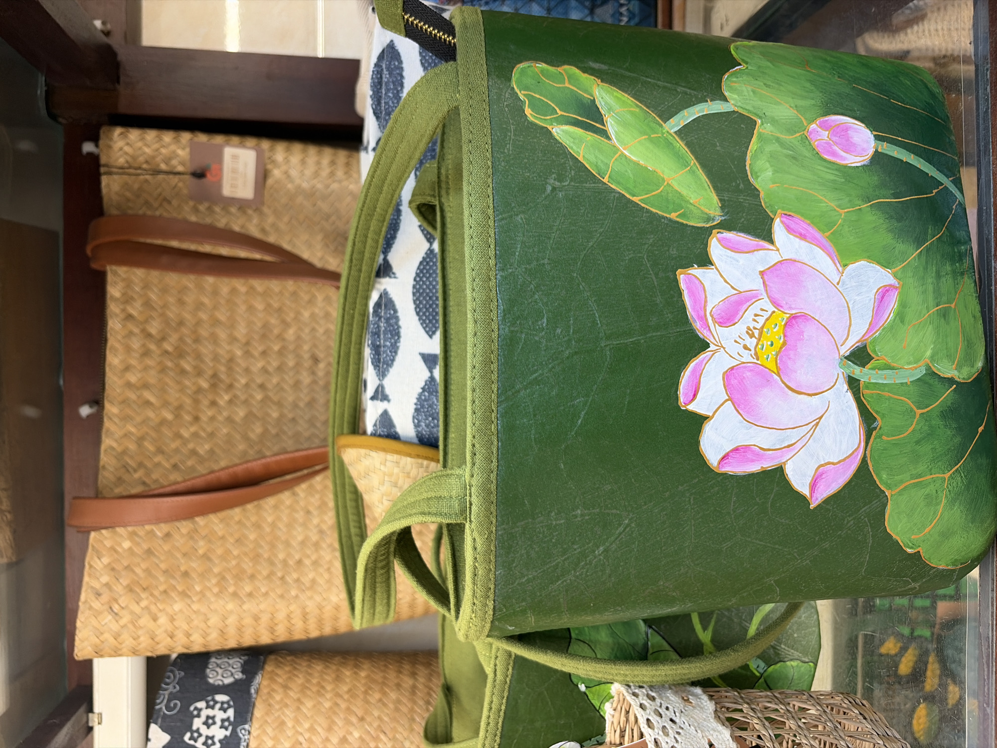 A close-up, eye-level, indoor shot captures a display of woven bags and a painted tote bag. In the foreground, a dark green tote bag with a painted lotus flower design dominates the frame. The bag has green fabric handles and trim. Behind it, a lighter beige woven tote bag with brown leather straps is partially visible. To the left of the woven tote, another woven basket with a white lace trim is seen. The background features shelves and other bags, suggesting a retail or market setting, possibly in Đà Nẵng, Vietnam. The lighting is moderate, highlighting the textures of the woven materials and the details of the painted design.