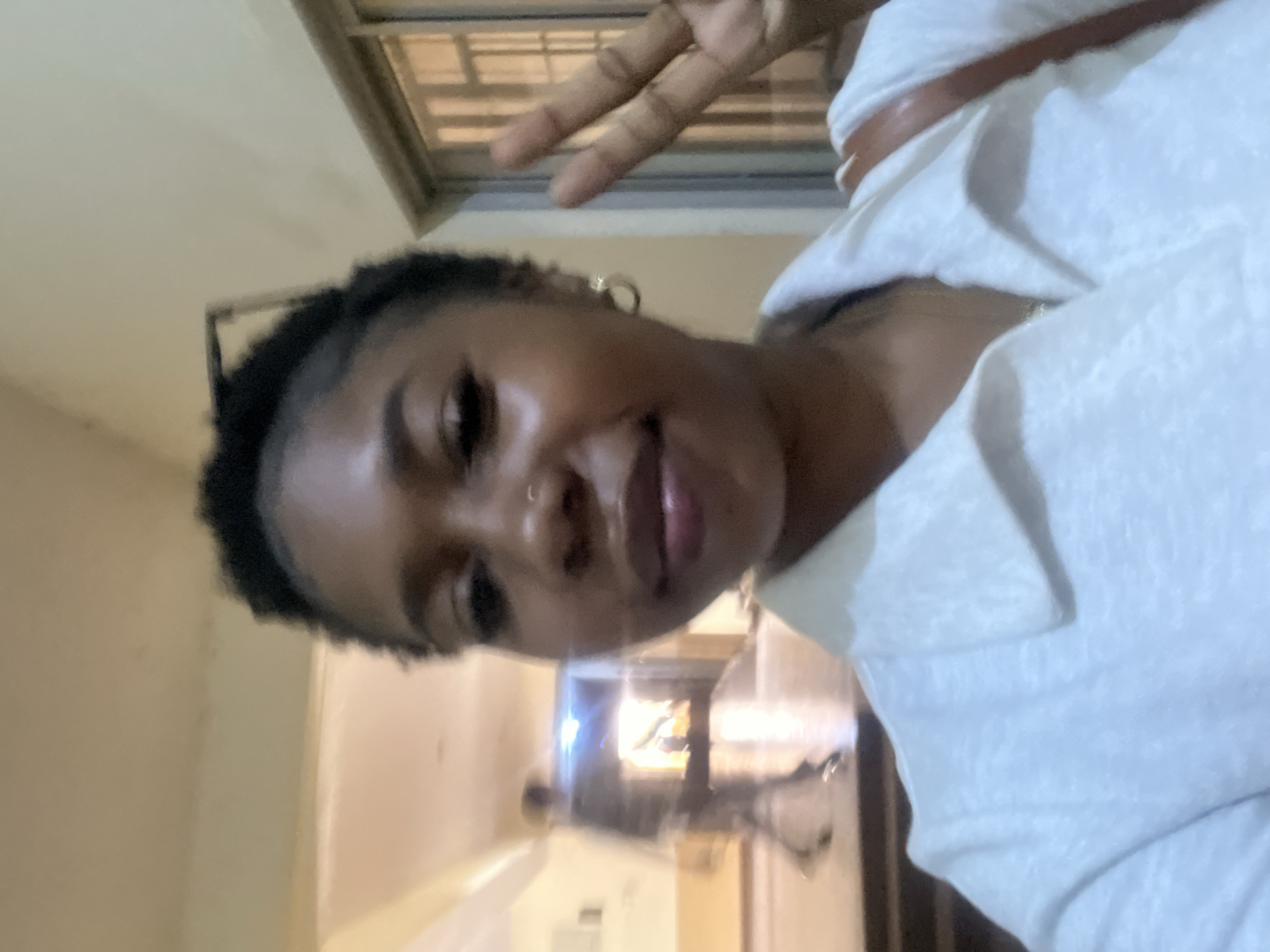In the foreground, a young Black woman, identified as being in Jos, Nigeria, takes a selfie. She is wearing a white collared shirt and has her hair styled in short dreadlocks. She is also wearing hoop earrings and a nose ring. She is holding up two fingers in a peace sign. In the background, a blurred figure of a person walks down a hallway. The hallway appears to be inside a building. There are windows on the right side of the hallway.