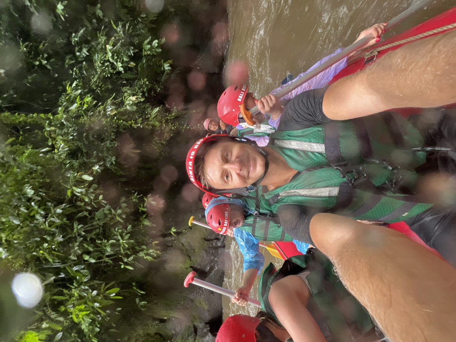 A group of people are white-water rafting. They wear helmets and life vests. The river's water is a murky brown. Lush green foliage borders the riverbank on one side. People in the raft are holding paddles. It's a close-up, possibly a selfie, with someone's arm slightly obscuring the foreground. Raindrops or water splashes are visible on the camera lens.