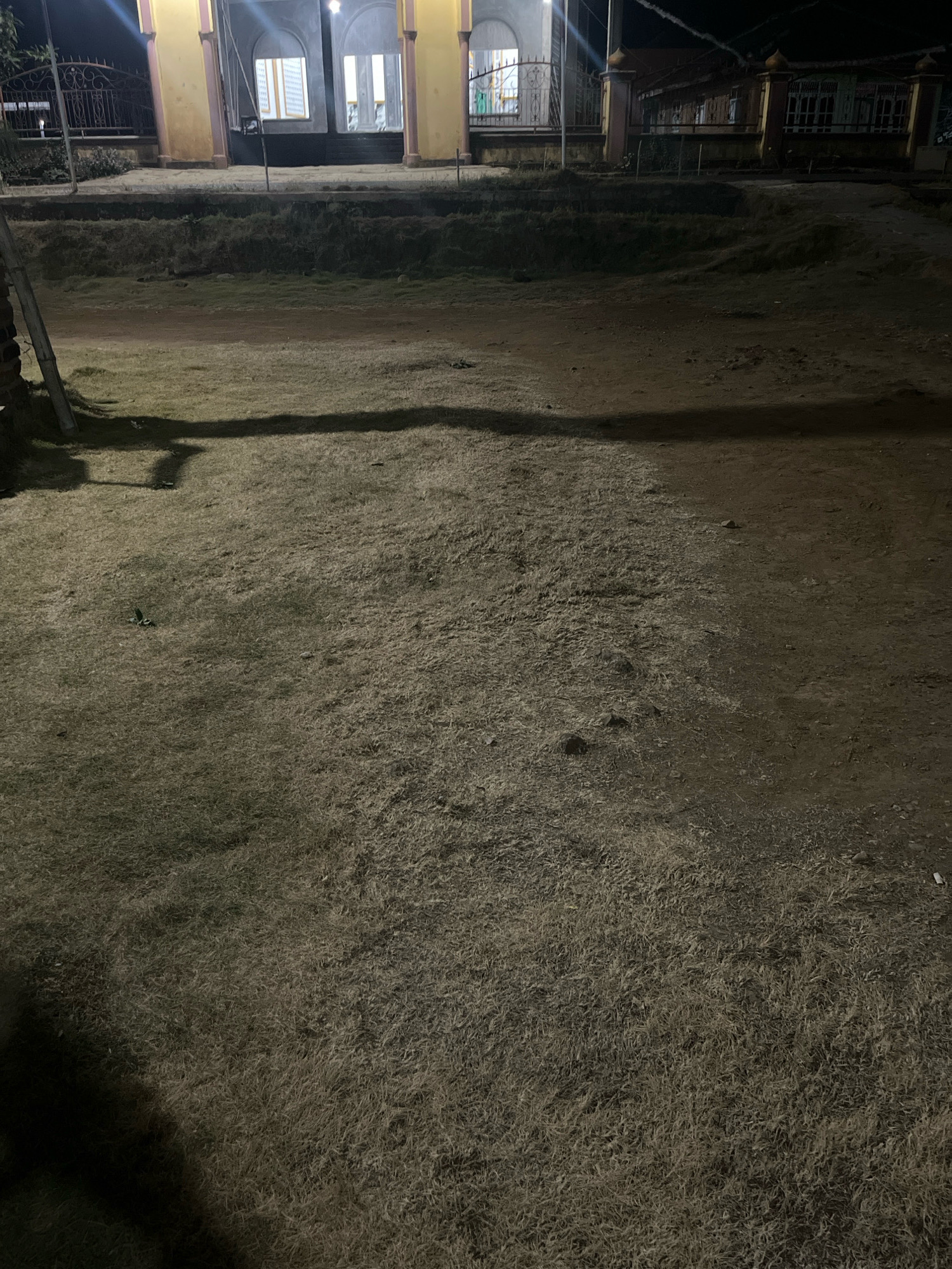 This image shows a nighttime view of a building with arched entrances and columns, possibly a place of worship or public building, situated in Kebumen, Indonesia. The building is illuminated from within, casting light onto the surrounding grounds. In front of the building is a flat area covered with dry, light-colored grass, which transitions into a dirt path. A shadow of a person is visible on the grassy area, suggesting someone was standing there or nearby when the photo was taken. A wooden pole is also present on the left side of the frame. The overall scene suggests a calm, perhaps deserted, environment at night.