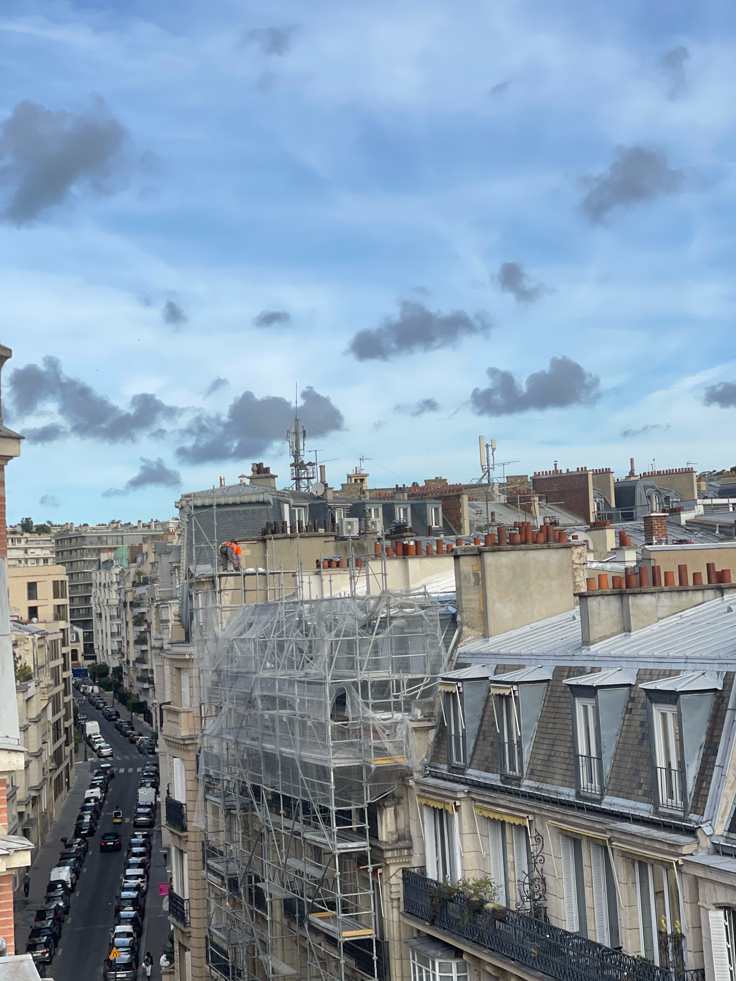 An elevated view captures a Parisian street scene under a cloudy sky. A narrow, tree-lined street is densely packed with parked cars, predominantly black and white, on both sides. Pedestrians are visible on the sidewalks, some walking, others standing. The buildings lining the street are typical of Parisian architecture, featuring multiple stories with mansard roofs, dormer windows, and decorative ironwork balconies. Scaffolding with white netting covers a building facade on the right side of the image, indicating ongoing construction or renovation. A construction worker in an orange vest is visible on the scaffolding. Rooftops with numerous chimneys are prominent in the midground, with telecommunication antennas perched atop some buildings. The sky is a mixture of blue and gray clouds, with a few small birds in flight.