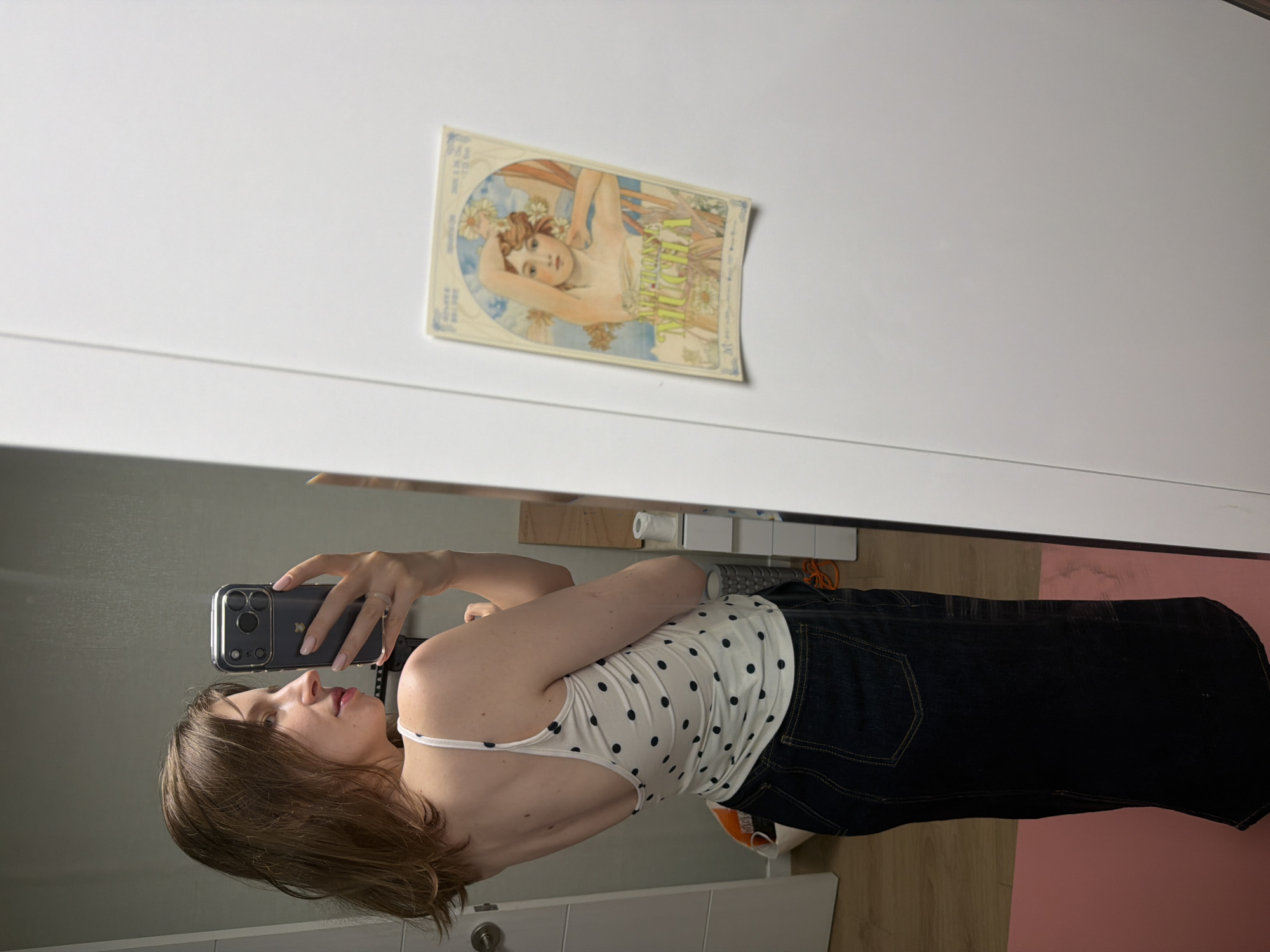A young woman stands in front of a full-length mirror, taking a selfie. She is wearing a white tank top with black polka dots and a dark denim skirt. Her hair is brown and shoulder-length. The mirror reflects her from the side, showing her face in profile as she looks at her phone. The background is a white wall, on which a decorative sticker of a woman with flowers is visible. A doorway with a white door is also visible on the left side of the frame. The floor is wooden, with a pink mat partially visible. The scene appears to be indoors, likely in a bedroom or dressing area. The image does not contain any specific landmarks or details that would allow for precise location identification within Dongducheon-si, South Korea.