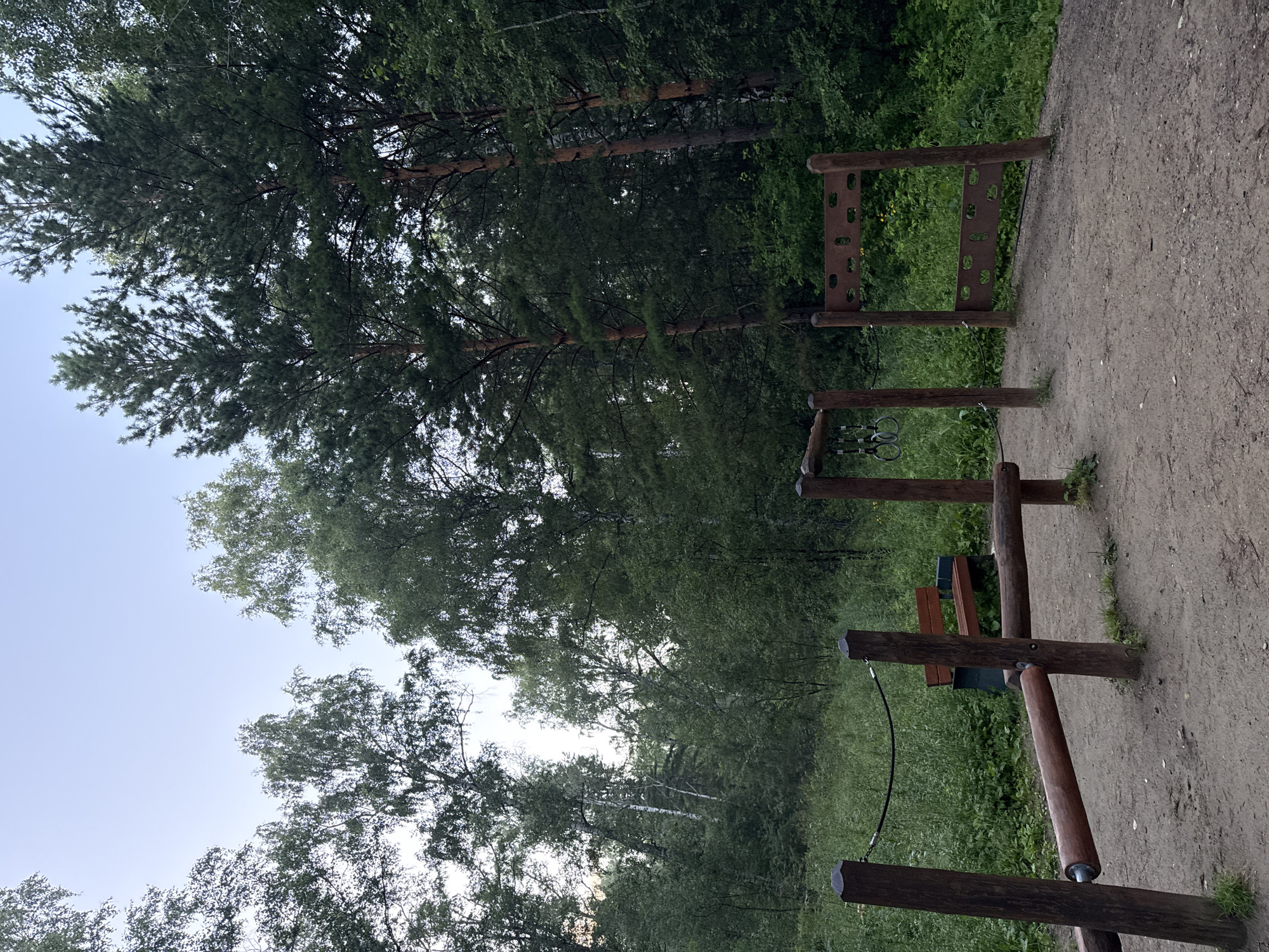A wooden outdoor gym set is visible in a wooded area. The gym equipment consists of several wooden posts supporting horizontal bars and a climbing structure. A wooden bench is situated behind the gym equipment. The ground is covered in dirt and scattered grass. In the background, there is dense foliage from trees and a clear sky above. The scene appears to be in a park or forest on the outskirts of Krasnoyarsk, Russia.