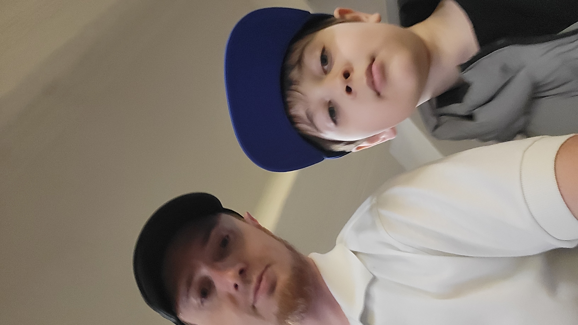 The image shows a close-up, low-angle shot of two people, an adult male and a young boy, both wearing baseball caps. The boy is positioned slightly above and to the right of the man. He is wearing a blue baseball cap and a dark shirt, and he is looking directly at the camera with a neutral expression. His face is partially obscured by the brim of his cap.
The man is wearing a black baseball cap and a light-colored collared shirt. His face is more visible, and he also appears to be looking towards the camera. He has a short beard and a serious expression. The background is a plain, light-colored wall or surface, providing no specific environmental clues. The lighting suggests an indoor setting.
The image is taken from a very low angle, as if the camera is looking up from the floor. This perspective makes the subjects appear larger and closer. The orientation of the image is also unusual, appearing to be rotated. There is no visible text in the image. The overall impression is that of a selfie or a candid shot taken by one of the subjects. The activity appears to be posing for a photograph.