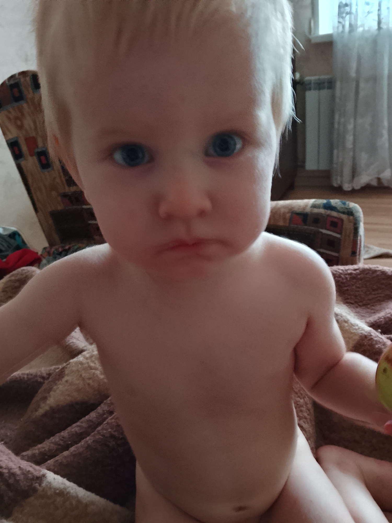 The media file is a close-up, low-angle, indoor shot of a young child, likely a toddler, with fair skin and light blonde hair. The child has bright blue eyes and is looking directly at the camera with a slightly pouty expression. They are bare-chested and appear to be sitting or lying on a brown and beige patterned blanket. In the background, a chair with a similar patterned upholstery is visible, along with a radiator and sheer curtains. The lighting suggests it might be daytime, but the overall atmosphere is intimate and personal, focusing solely on the child. There is no visible text in the image. The provided location context places the scene in Kaluga, Russia.