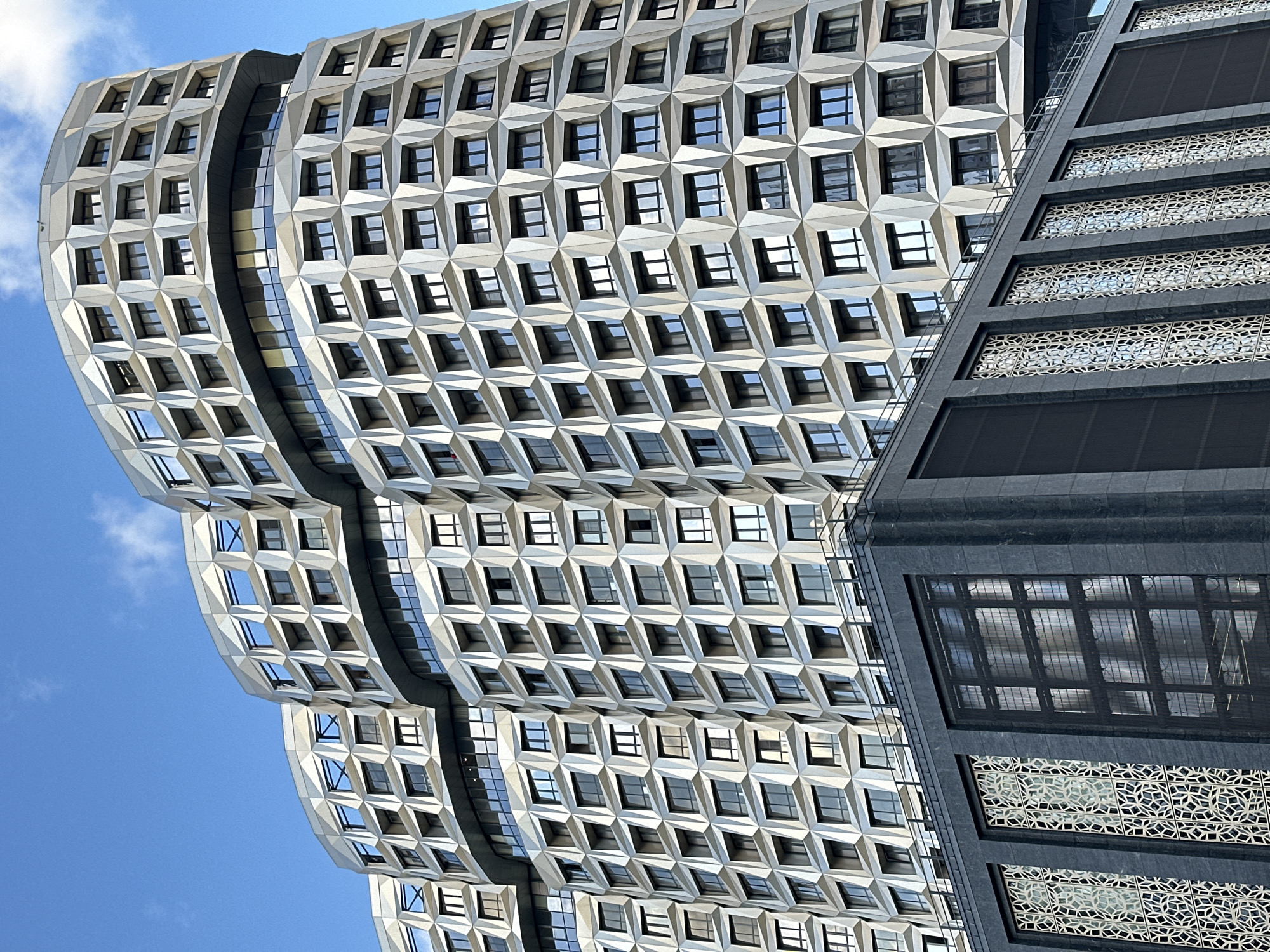 This is a low-angle, full-shot photograph of the white geometric facade of a modern building in Moscow, Russia. The building is comprised of multiple curved sections, each featuring a repeating pattern of diamond-shaped recesses housing dark-paned windows. The facade itself appears to be a metallic material that reflects the blue sky. At the base of this structure is a darker, lower building with a granite-like facade adorned with intricate, gold-colored geometric patterns on some of its windows. The sky is clear blue with a few faint white clouds visible. The scene is devoid of any people or visible activity, presenting a clean, architectural study.