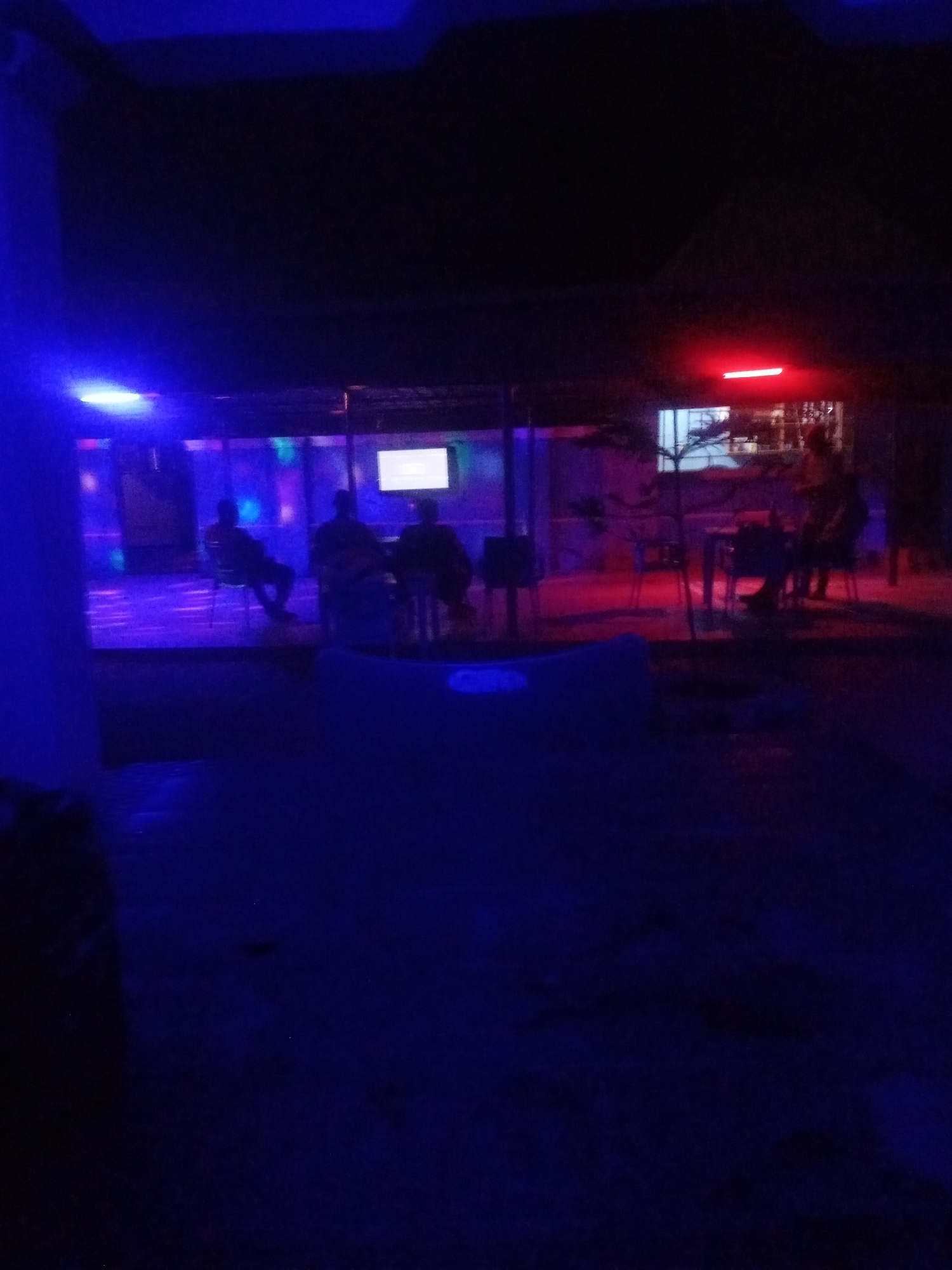 This is a nighttime photograph taken at the Blue Sea Bar and Restaurant in Sabon Gari, Nassarawa, Nigeria. The scene is illuminated by a combination of blue and red lights. In the background, several patrons are seated at tables, and a television screen is visible. Some of the patrons appear to be engaged in conversation, while others are observing the screen. The establishment has an open-air or semi-open-air seating area, with a roof structure visible in the upper portion of the image. The overall atmosphere suggests a lively social gathering or entertainment venue.