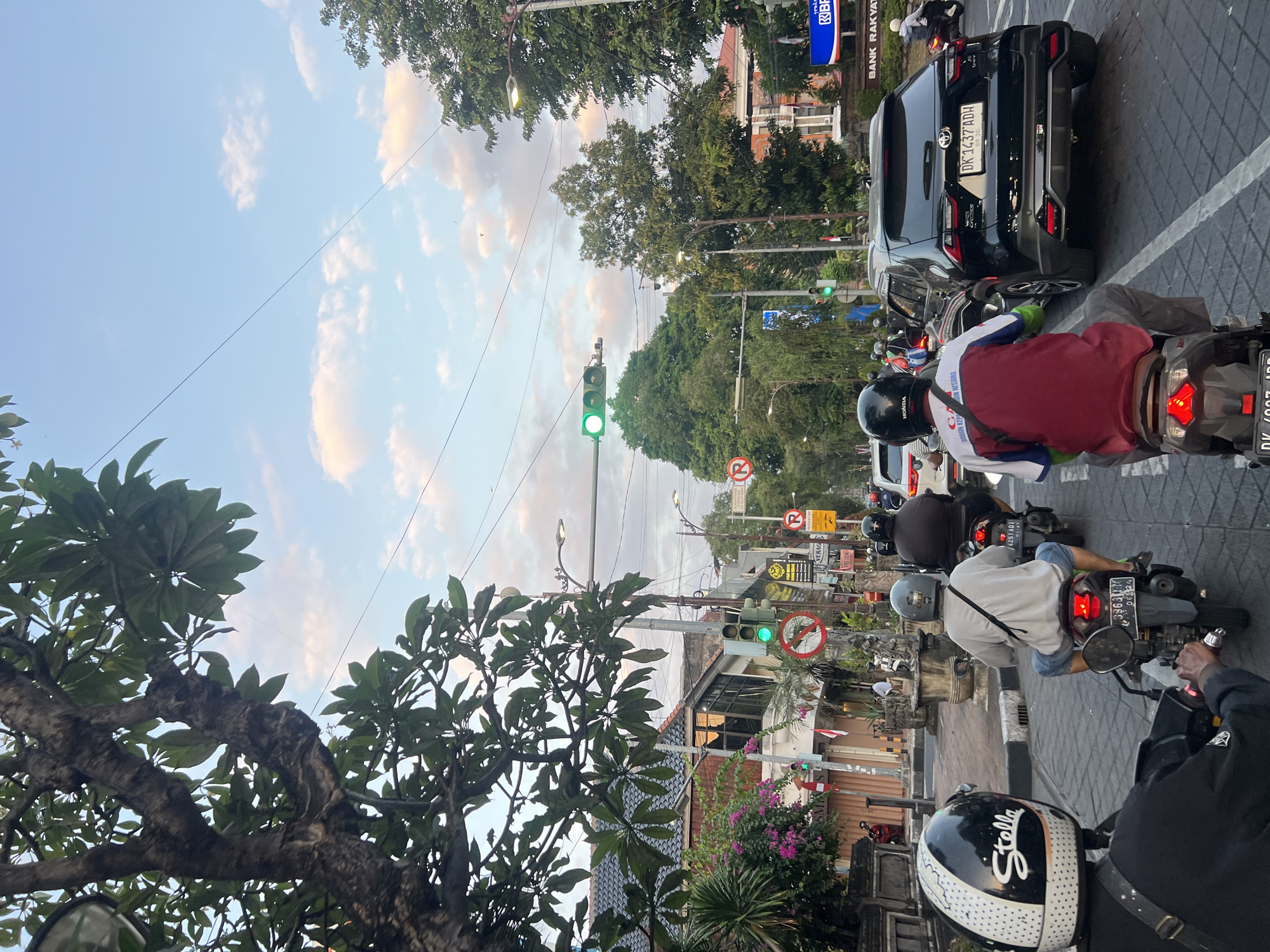 The image depicts a street scene in Denpasar, Indonesia, likely during the late afternoon or early evening, given the lighting and sky. The perspective is from the back of a motorcycle, looking forward through traffic. In the foreground, the back of a motorcyclist wearing a black helmet with a white "Stella" logo is visible. To their right, another motorcyclist in a grey t-shirt and blue shorts is also on a motorcycle. Further ahead, a black SUV is stopped, and beyond that, more motorcycles and cars are in traffic.
The street is lined with buildings, some appearing to be shops or businesses, with awnings and signage. There are traffic lights, one of which is illuminated green. Circular red and white traffic signs are also visible on poles. Lush green trees line parts of the street, and overhead, power lines crisscross the sky, which is a pale blue with scattered white clouds tinged with pink. The overall impression is a typical urban traffic scene in a tropical environment.