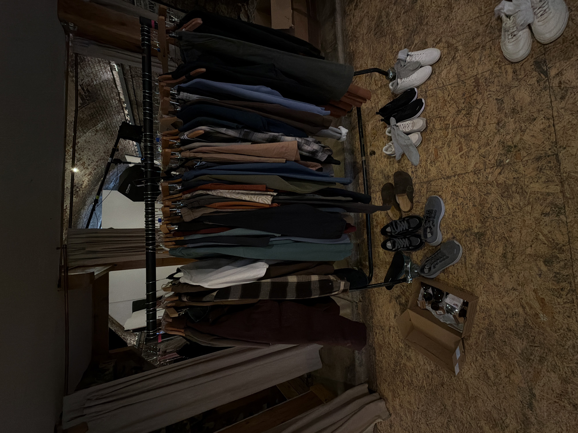 A cluttered interior is visible. A metal clothing rack filled with clothes dominates the view. Several pairs of shoes are scattered on the wooden floor. A cardboard box sits on the floor with smaller items inside. The setting appears to be a room within a building. There is no indication of it being in the city of London, United Kingdom. No people are visible.