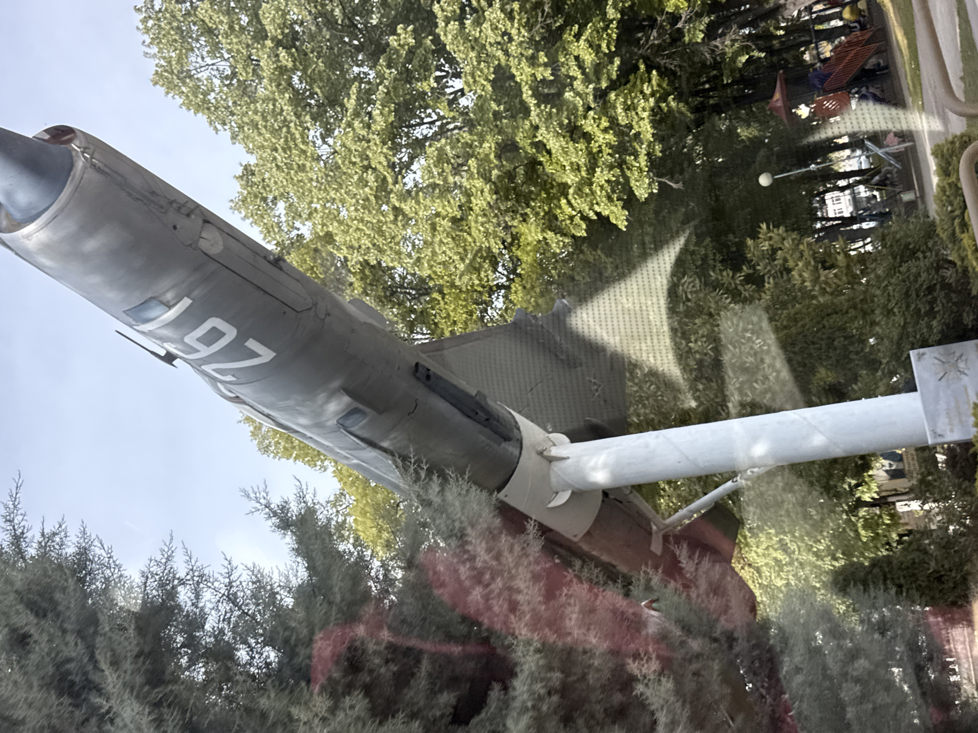 The image depicts a MiG-21 fighter jet mounted on a pedestal in the village of Yoakim Gruevo, Bulgaria. The aircraft is painted in a gray color scheme with the number "92" clearly visible on its fuselage. The jet is positioned at an upward angle, suggesting a symbolic representation of flight. The surrounding environment includes mature trees and a glimpse of what appears to be a park or recreational area in the background. The photograph seems to be taken through a window, indicated by reflections on the glass. A white support pole is holding the aircraft. Vegetation is visible both in front of and behind the aircraft.