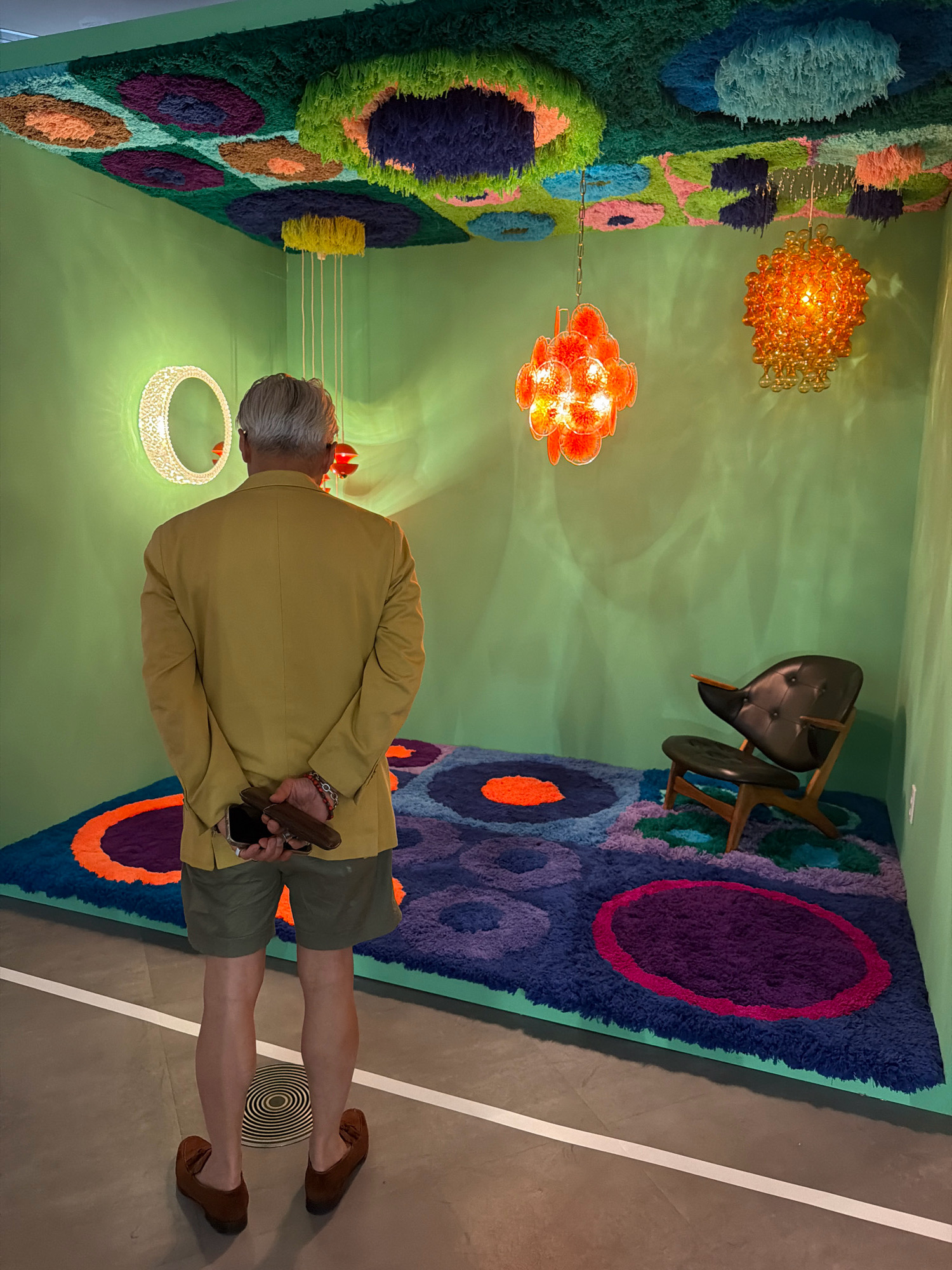 A man stands in a room with bright green walls, facing away from the camera. He has gray hair and is wearing a yellow blazer over olive green shorts. His hands are clasped behind his back, holding a phone and a dark brown object. On the floor, a plush rug with circular patterns in blue, purple, and orange is laid out. A dark leather chair with wooden legs sits in the corner. The ceiling is covered with colorful, shaggy, textured panels in shades of green, blue, pink, and purple. Several decorative light fixtures hang from the ceiling, including a circular, frosted white one on the left wall and two orange, orb-like chandeliers. The room is brightly lit, with shadows cast on the walls. This scene is likely from an art installation or exhibition, potentially at a gallery or museum in Seoul, South Korea.
