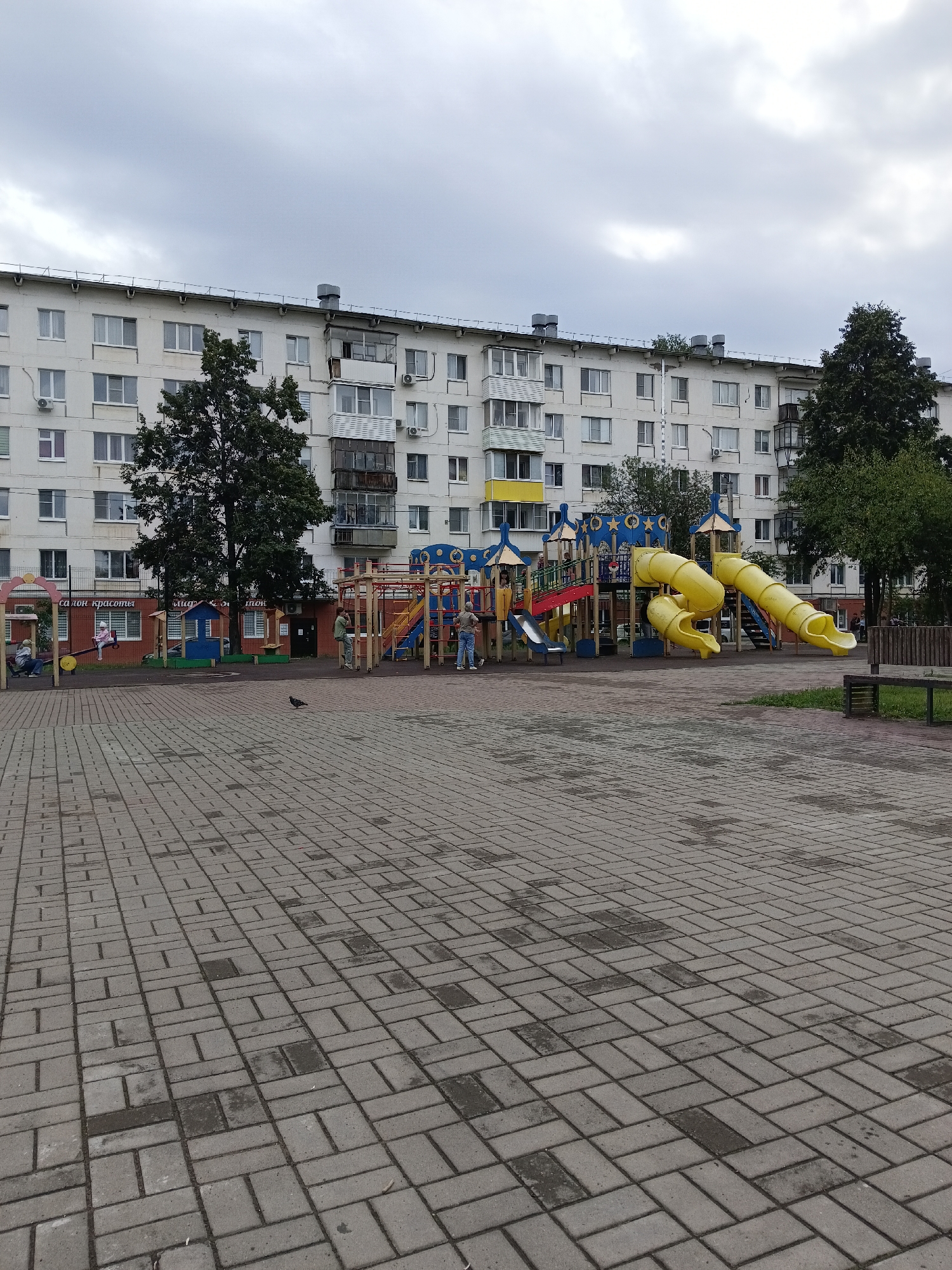 A wide shot captures an overcast day in Chaikovsky, Russia, with a multi-story residential building dominating the background. The building is characterized by its white facade, numerous windows, and visible air conditioning units. In the foreground, a paved courtyard features a children's playground with bright yellow and blue structures, including slides and climbing equipment. A few individuals are present in the scene: a person is seen on a swing, another stands near the playground, and a third is walking on the paved area. A pigeon is also visible on the ground. The overall atmosphere is subdued, reflecting the cloudy weather.