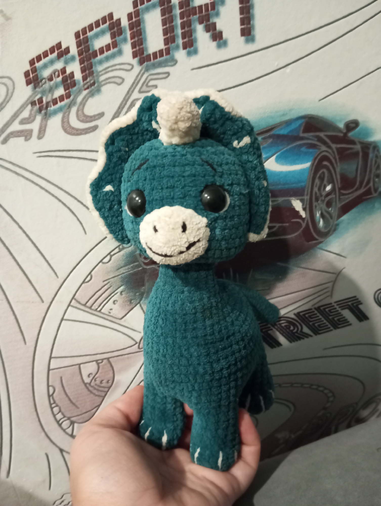 A crocheted toy dinosaur with teal-colored yarn and white accents is being held by a person's hand against a backdrop of a wall featuring graphics of a blue sports car and lettering that reads "SPORT" and "STREET". The toy has large black eyes, a white snout with a smiling mouth, and two small white tufts on its head. The wall has a repetitive geometric pattern in a light gray color. The scene appears to be indoors. No specific location in Verkhnyaya Pyshma, Russia can be identified from the image.