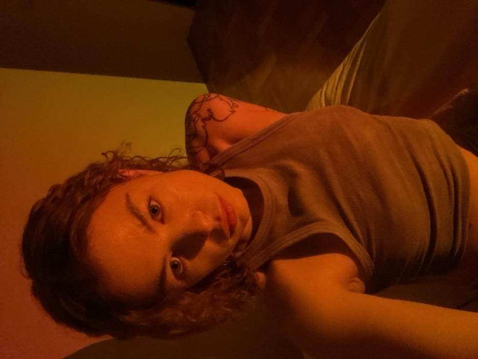 A woman lies on her side, facing the camera. Her hair is curly and brown. She wears a gray tank top, exposing her shoulder and a tattoo. The lighting casts an orange hue across her face and body. She has light eyes and pink lips. The background is blurry, but appears to be a wall and soft furniture, also bathed in the warm, orange light.