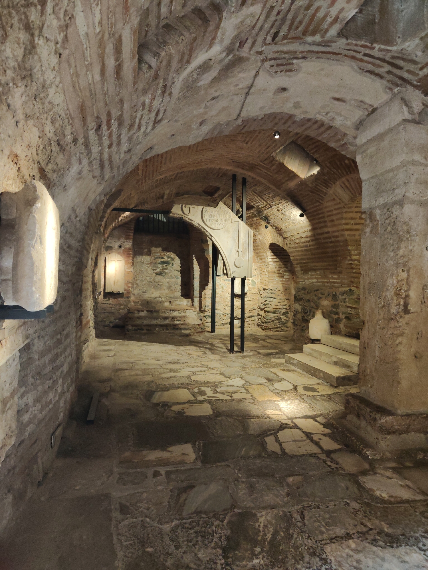 The image depicts the interior of the Rotunda of Galerius in Thessaloniki, Greece. It is a dimly lit, ancient stone passageway with a vaulted brick ceiling. Large, irregular flagstones pave the floor, some appearing wet and reflecting the light from strategically placed spotlights.
On the left, a large, carved stone fragment, possibly a column capital or a piece of architectural decoration, is mounted on a dark metal stand. Further down the corridor, a similar, but smaller, stone artifact hangs from black metal supports. This artifact appears to have some inscriptions on it.
The walls are constructed of rough-hewn stone and brickwork, showing signs of age and wear. In the background, there are more arched openings and steps leading up to an elevated area. A white ceramic amphora sits on these steps, illuminated by a spotlight. Several spotlights are visible throughout the passageway, casting pools of light and shadows, highlighting the textures of the stone and brick. The overall atmosphere is one of historical preservation and archaeological interest.
