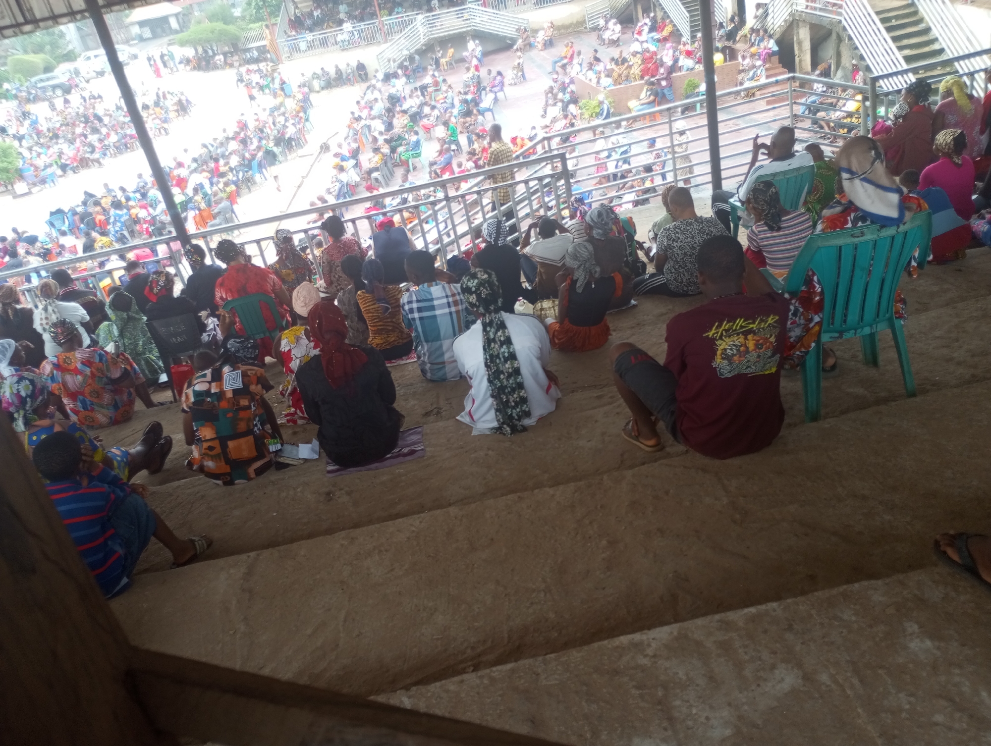 A medium shot captures a group of people sitting on concrete steps in an outdoor setting, likely a public gathering space or an event venue, possibly in Onitsha, Nigeria. The majority of the individuals are seated, facing away from the camera and towards something out of frame, suggesting they are attending a performance, religious service, or a public address. Their attire is diverse, with many wearing brightly colored fabrics, typical of Nigerian fashion. Some are wearing headwraps or scarves. Several individuals are sitting on the ground or on small mats, while others are seated on plastic chairs, some of which are a distinctive teal color.
In the background, a larger crowd of people is visible, some standing and some seated in rows on elevated platforms or bleachers, also facing the same direction. The architecture of the background suggests a multi-tiered structure with metal railings, resembling a stadium or an auditorium. There are also trees and some distant buildings visible, indicating an urban or semi-urban environment. The overall atmosphere appears to be one of a communal event, with a focus of attention directed away from the viewer. The lighting suggests it is daytime, possibly overcast or under a covered structure, as the shadows are not harsh. The scene is captured from a slightly elevated angle, looking down upon the group in the foreground.