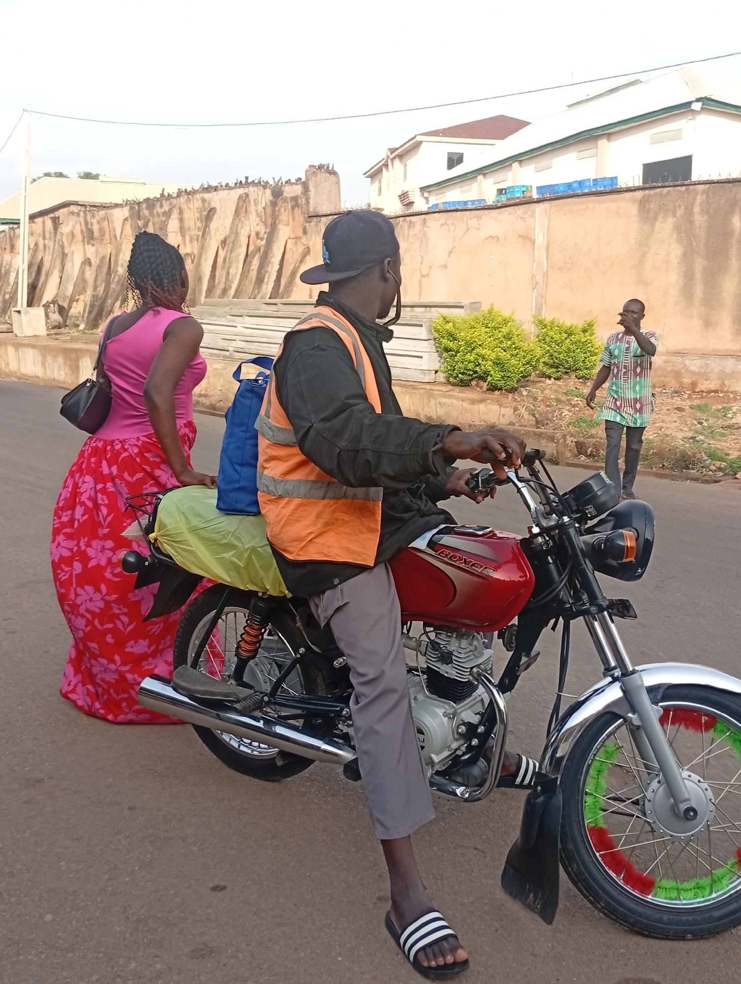 A motorcylist sits on a red "Boxer" brand motorcycle in Gura Top, Nigeria. The rider, wearing a backwards black cap, black jacket, and orange safety vest, is positioned on the road, ready to depart. A woman in a pink top and red floral skirt stands beside the motorcycle. A blue bag sits on the back of the motorcyle behind her, covered in yellow plastic. Another man in a patterned shirt is walking down the road in the background. A stone wall, bushes, and buildings with brown roofs are visible in the background.