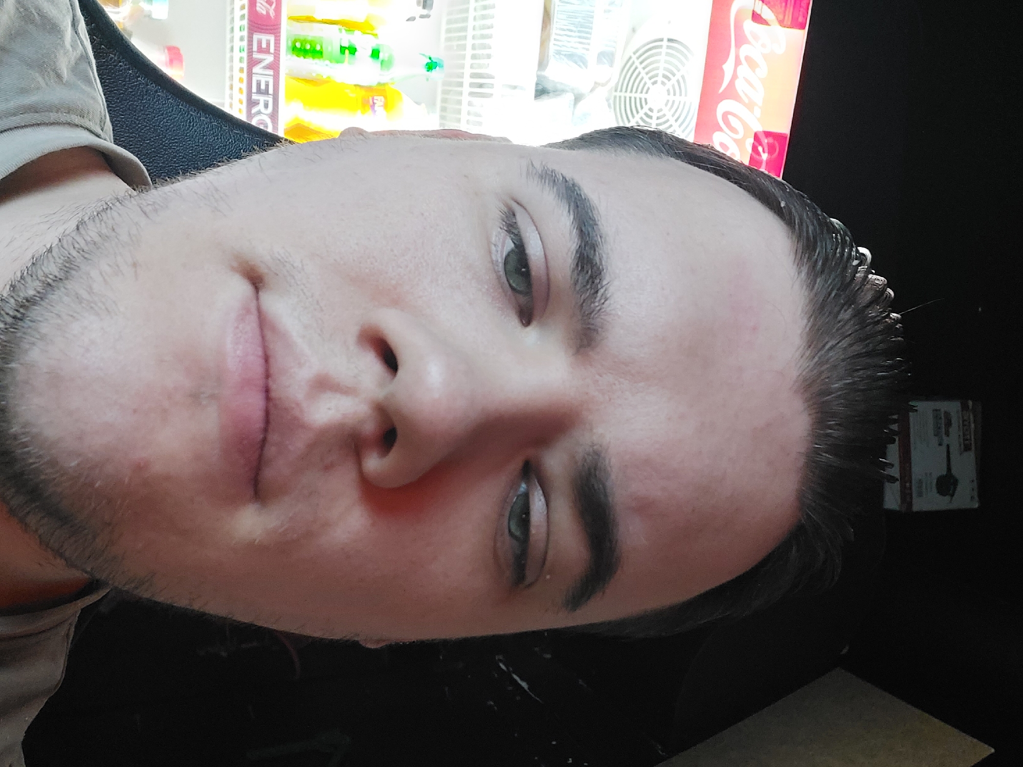 A close-up photo of a man's face, slightly angled. The man has fair skin, dark eyebrows, and green eyes. He has short, dark hair that appears to be slicked back. He also has stubble. He is positioned in front of a vending machine. The vending machine has a Coca-Cola sign on it and contains various beverages and snacks. The location is in Tashkent, Uzbekistan.