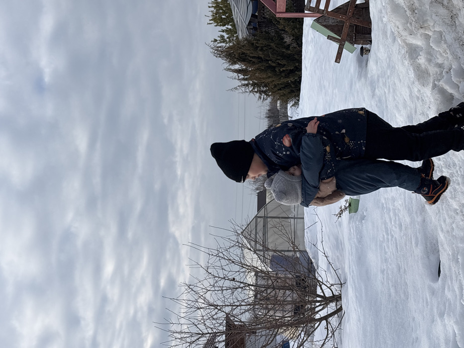 Two individuals are depicted outdoors in a snow-covered environment under an overcast sky in the city of Olkhovka, Russia. The taller individual, standing on the right, wears a black beanie and a dark jacket with light-colored celestial patterns, embracing a smaller individual. The smaller individual, wearing a light gray knitted beanie with a pompom and a dark snowsuit, is held off the ground by the taller person, with their head resting against the taller person's shoulder. In the background, to the left, a bare deciduous tree stands in front of a translucent, arched greenhouse structure. To the right, a cluster of dark green coniferous trees is visible, along with the partial view of a small, rustic wooden windmill-shaped ornament. The ground is uniformly covered in snow.
