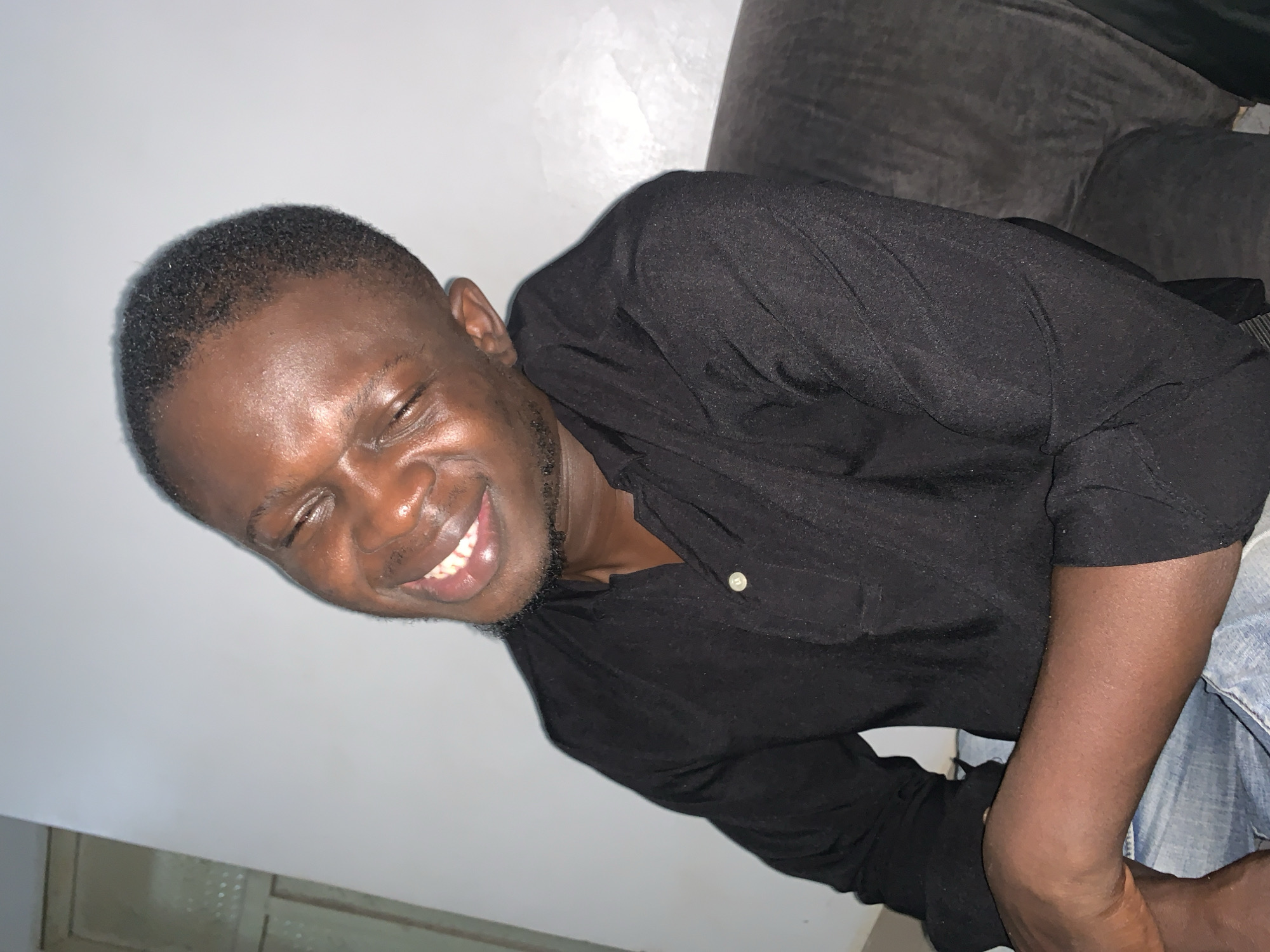 A young Black man with his eyes closed and a wide smile is captured in this medium shot. He is leaning back with his head tilted to his left, and his hands are clasped. He is wearing a black button-up shirt with a collared neckline and light blue jeans. The background is a plain, light gray wall. The scene appears to be indoors, possibly in a residential setting in Abuja, Nigeria, with the lighting suggesting an evening or nighttime capture.