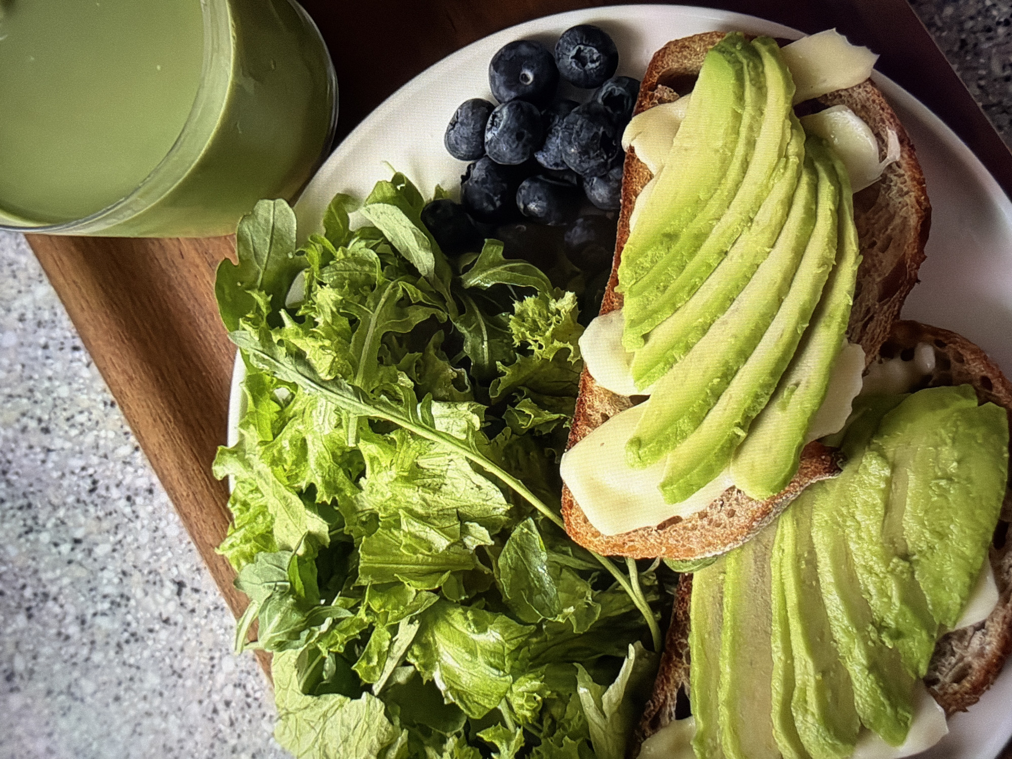 The image displays a close-up, overhead view of a healthy meal set indoors, likely in Ros Na Rí, Ireland. On the left, a clear glass filled with a vibrant light green smoothie or juice rests on a wooden surface. A white, oval-shaped plate dominates the center, holding various food items.
On the plate, a large serving of fresh green salad, appearing to be a mix of lettuce and arugula, occupies the lower left section. Adjacent to the salad, a small pile of dark blue blueberries adds a fruity element. The primary components of the meal are two slices of what appears to be whole-grain toast. Each toast slice is generously topped with creamy white cheese, possibly melted, and meticulously fanned slices of bright green avocado.
The plate and glass are positioned on a warm-toned wooden board or tray, which itself is resting on a light-colored, speckled countertop or table. The lighting suggests daytime, enhancing the natural colors of the fresh ingredients. There are no people visible, nor any discernible text in the frame. The overall scene conveys a focus on fresh, nutritious eating with a clean and appealing presentation.