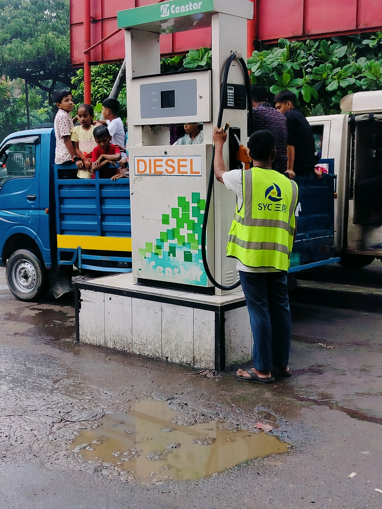 In Dhaka, Bangladesh, at a petrol station, a blue truck is parked next to a diesel pump. Several children are in the back of the truck, some standing and some sitting. A man wearing a high-visibility vest is operating the diesel pump. The ground is wet and muddy with several puddles. The petrol station appears to be an outdoor facility with some greenery visible in the background.