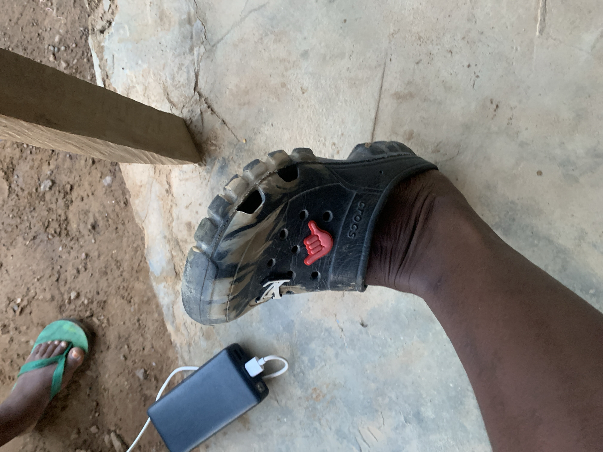 This close-up, high-angle photograph captures a ground-level scene in Keffi, Nigeria, featuring a person's leg and footwear, a power bank, and another person's foot.
The primary focus is a dark-skinned leg and foot, likely a right leg, wearing a black Croc shoe. The Croc appears worn and covered in dried mud or dirt, and features visible "Crocs" branding on its side. Two decorative Jibbitz charms are inserted into the shoe's ventilation holes: a prominent red "shaka" or "hang loose" hand gesture, and a smaller white charm that resembles a stylized letter "A" or a logo.
Adjacent to the Croc, a black power bank with a white charging cable is positioned on the ground, suggesting a device, not visible in the frame, is being charged.
To the left of the power bank, a portion of another dark-skinned foot is visible, wearing a bright green flip-flop or sandal.
The ground surface is a blend of light grey, cracked concrete or cement in the foreground and a brownish-orange, dusty dirt or sandy area with small stones towards the background. A rough wooden plank or beam extends diagonally across the upper left portion of the image.
The scene suggests an outdoor or semi-outdoor, informal setting, typical of a warm climate. The lighting is bright and even, indicating it is daytime. No specific emotions are discernible. The only visible text is "Crocs" on the shoe.