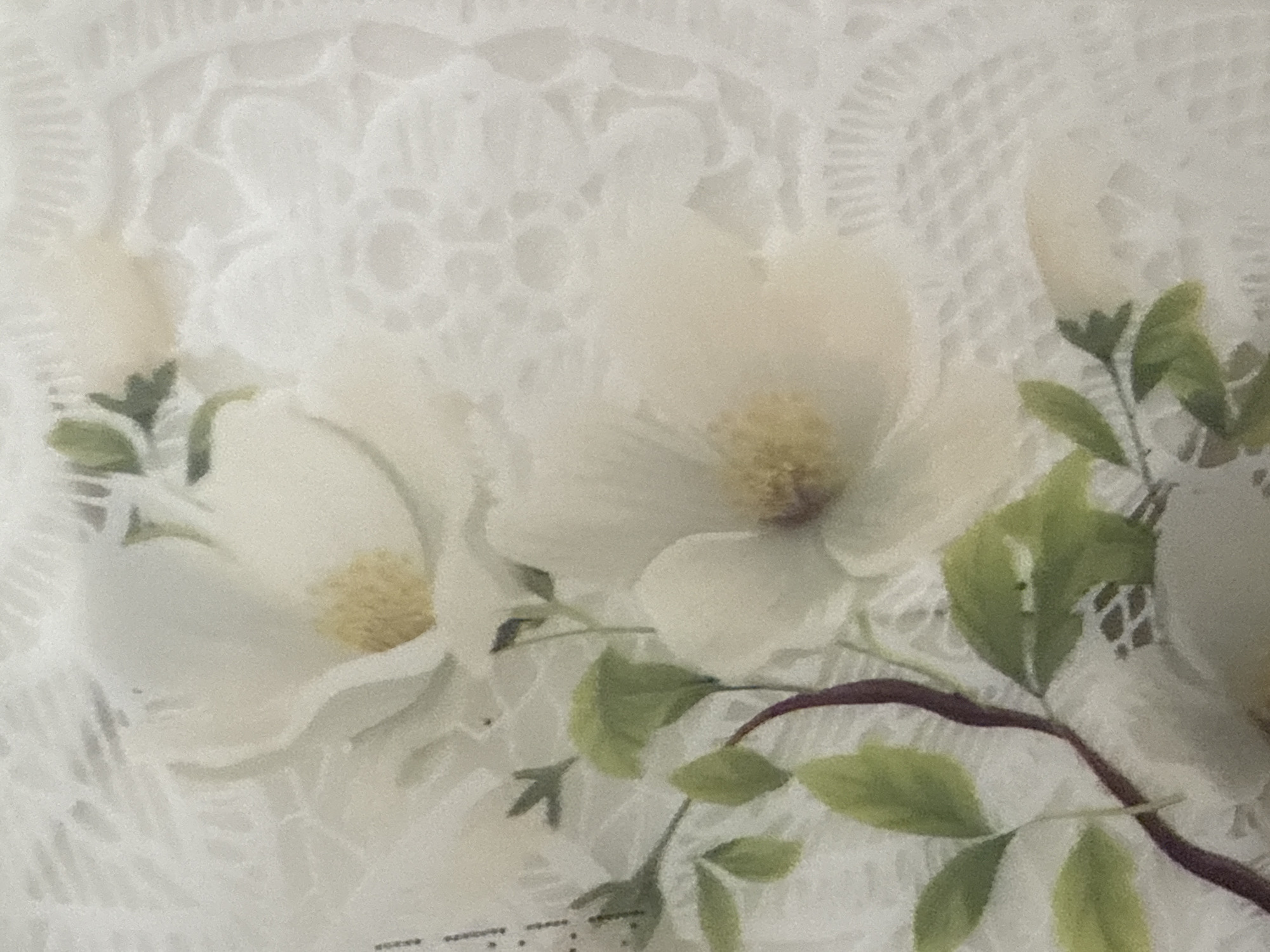 The image shows a close-up of a white floral pattern on a lace-like background. The flowers are rendered in soft, painterly strokes, with subtle yellow centers. Green leaves and a dark, winding stem are also depicted, adding depth and contrast to the composition. The overall impression is delicate and elegant. The image is a photograph of a textile, possibly a tablecloth, curtain, or piece of clothing. There is no information in the image to indicate its location in the city of Ushtobe, Kazakhstan.