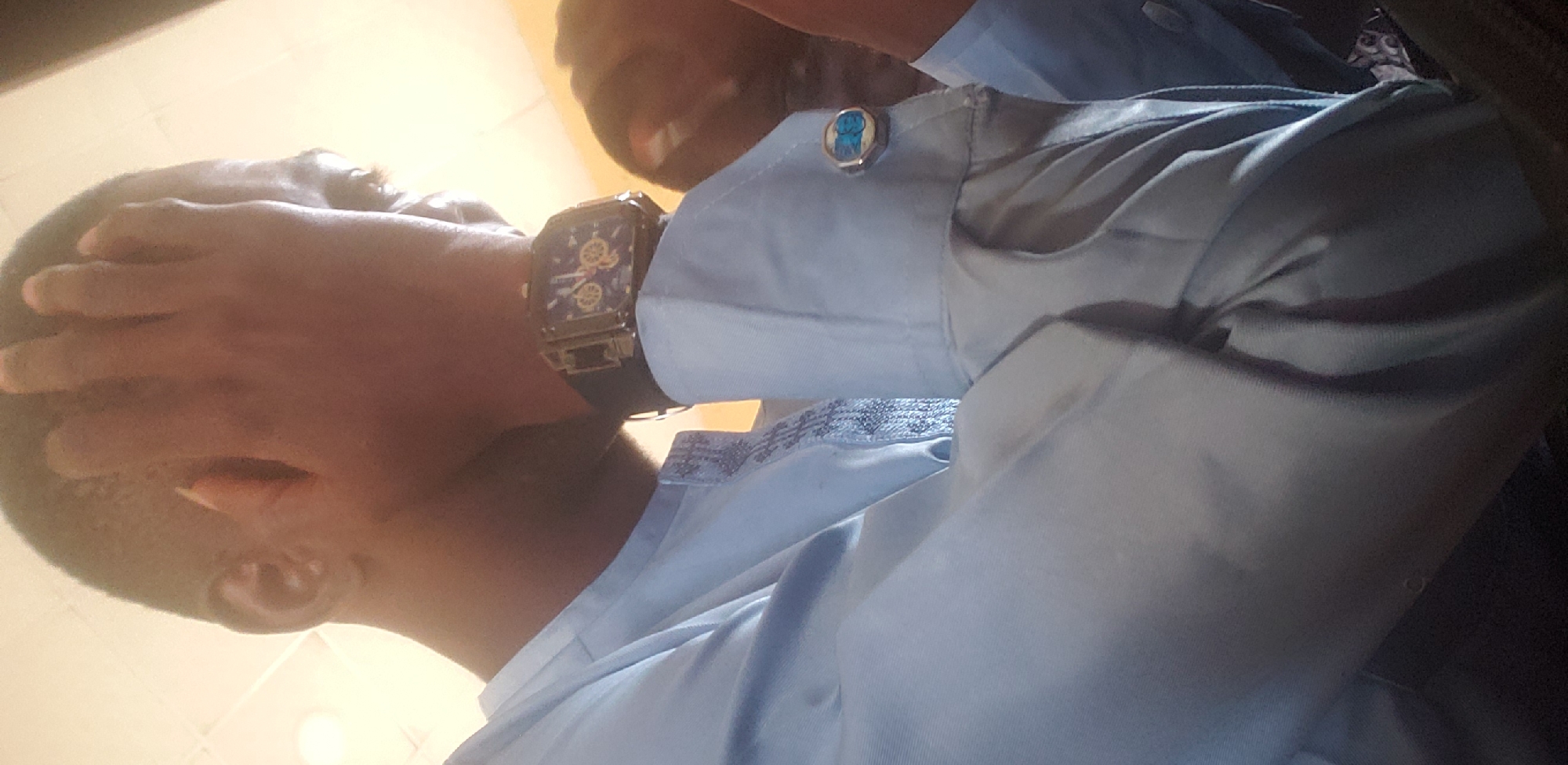The image shows a close-up of a man's head and upper body, seen from a low angle. His right hand is covering his face. He is wearing a light blue collared shirt with patterned trim on the placket and a watch on his left wrist. A cufflink with a blue stone is visible on his left shirt cuff. The background is out of focus, but appears to be an indoor setting with a light-colored ceiling. Another person's head is visible in the background on the right side of the image. The overall impression is that the man is either shielding himself or expressing distress.