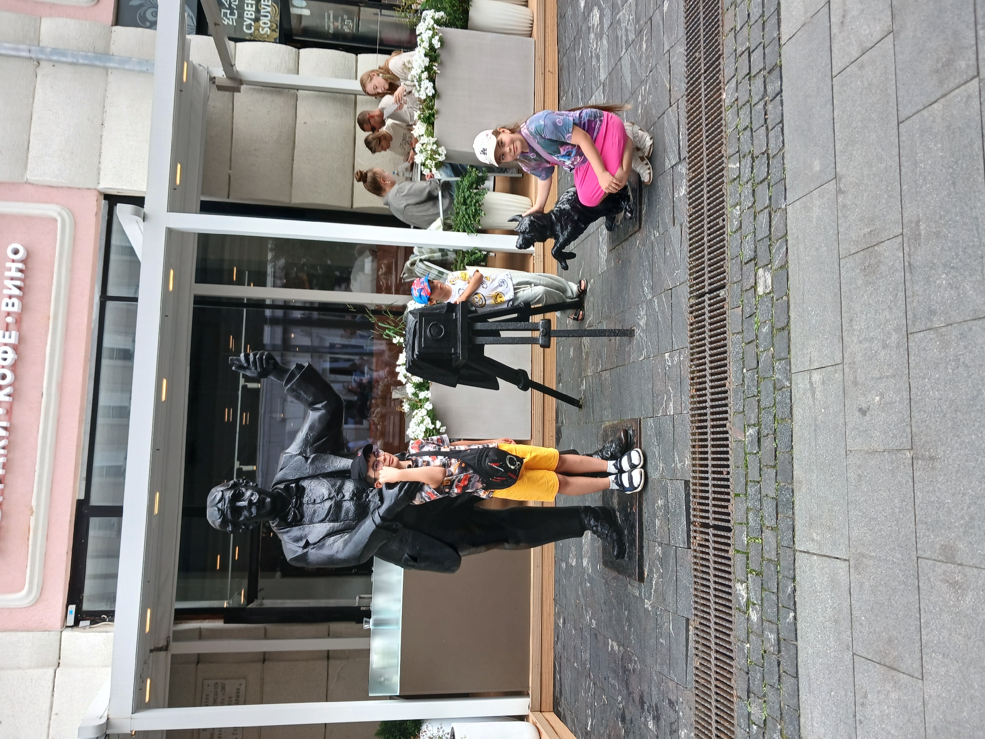 In Nizhny Novgorod, Russia, a bronze statue of a man holding a camera stands on a cobbled plaza in front of a building with a pink facade. The statue is positioned near a black metal grate covering a drain. To the left of the statue, a young boy in a patterned t-shirt and yellow shorts stands next to the statue's base, holding a black bag. To the right and slightly in front of the statue, a young girl in a colorful t-shirt and pink skirt is crouching next to a black statue of a dog. Behind the girl and the dog, another young boy wearing a baseball cap and a t-shirt stands near a vintage camera on a tripod. In the background, several people are seated at tables outside a cafe. The overall scene depicts a public outdoor space with art installations and people enjoying the environment.