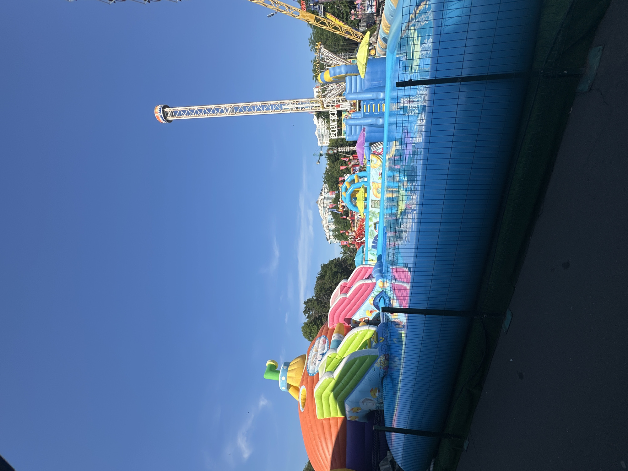 A bright, sunny day at an amusement park is depicted. The foreground features a large, inflatable water play area with a blue pool and various colorful slides and structures, including a large orange one with green and pink slides. The water is reflecting the vibrant colors of the inflatable toys. A black metal fence encloses the water area, and a dark paved path runs alongside it. In the background, a tall, yellow amusement park tower with a red and white striped top stands prominently against a clear blue sky. Other amusement park rides, such as a yellow roller coaster structure, are visible further in the distance, along with buildings and trees. The overall scene is lively and suggests a fun outdoor recreational environment.
