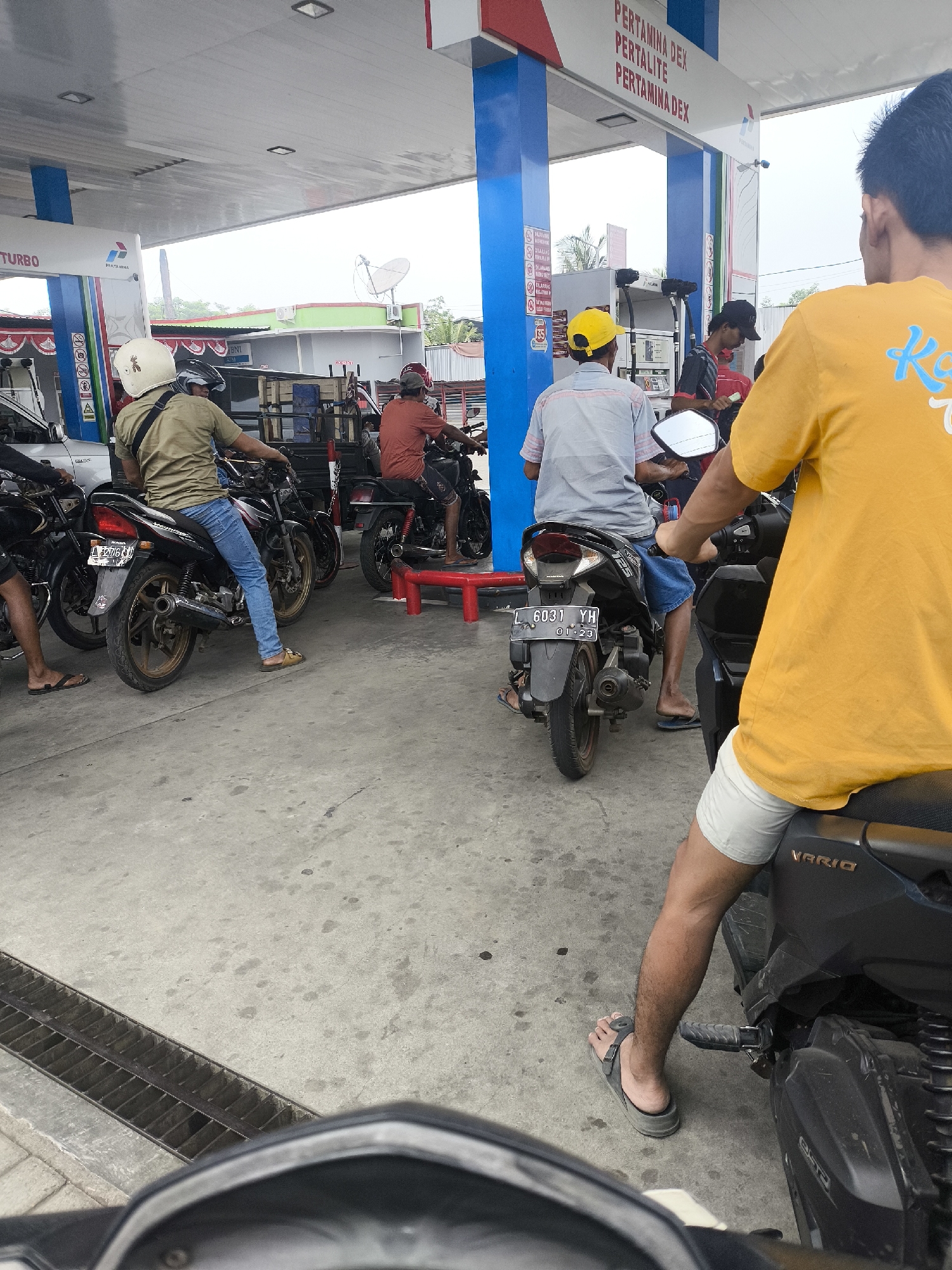 The image depicts a scene at a Pertamina gas station in Purworejo, Indonesia. Several individuals on motorcycles are queuing to refuel. The gas station features a blue and white canopy supported by blue pillars. Visible signage on the canopy includes "PERTAMINA DEX," "PERTALITE," and "PERTAMINA DEX."
In the foreground, a person wearing a yellow t-shirt and white shorts is seated on a black motorcycle, facing away from the camera. To their left, another person in a grey and white striped shirt and blue shorts is also on a motorcycle. Further left, a person in a khaki shirt and blue jeans is on a black motorcycle, with their left leg extended.
In the middle ground, a person in an orange t-shirt is seated on a red motorcycle. Behind them, another person in a red shirt and black shorts is also on a motorcycle. The ground is concrete, with a metal grate visible in the lower left corner. The background shows various buildings and structures, including a satellite dish on a rooftop. The overall atmosphere suggests a typical day at a busy gas station.