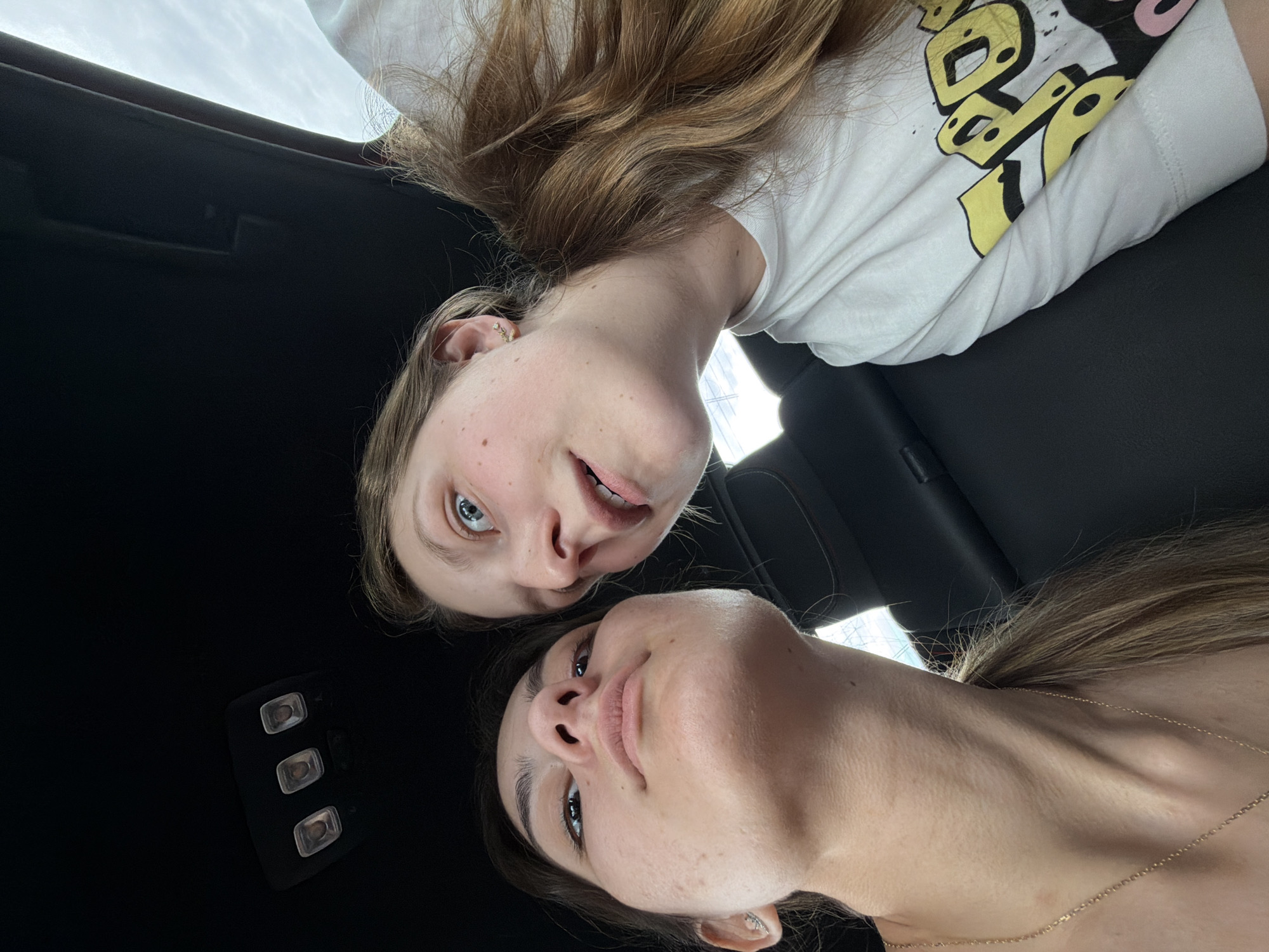 The image shows two women looking at the camera, appearing to be in a vehicle. The woman at the top of the image has lighter hair and is wearing a white t-shirt. The woman at the bottom has darker hair and is wearing a delicate necklace. The interior of the vehicle is visible, showing a black ceiling and seat. The location is not discernible from the image.