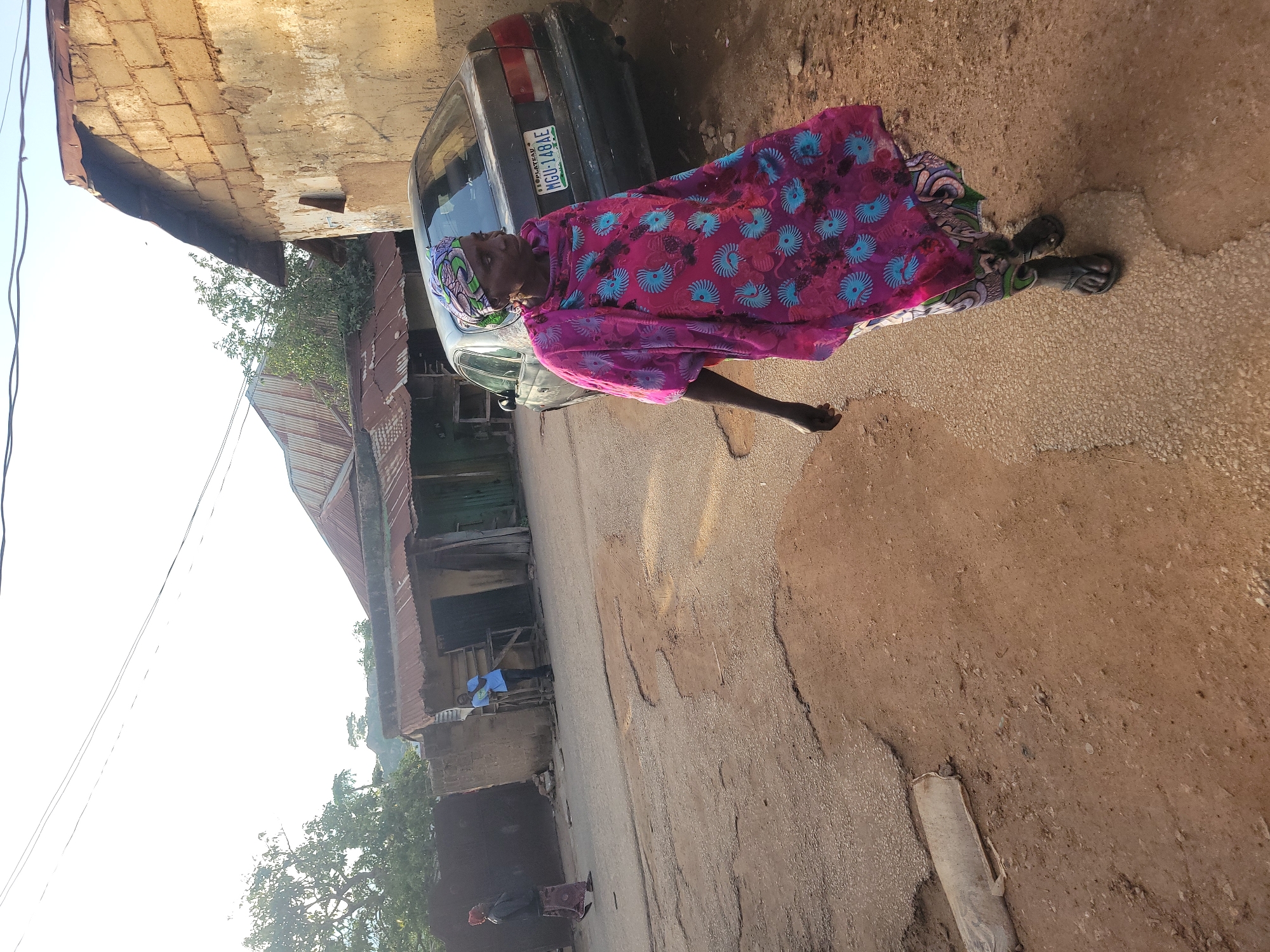 This image captures an outdoor street scene in the city of Jos, Plateau State, Nigeria, under bright, slightly hazy daylight. In the immediate foreground, an adult female with dark skin walks towards the right of the frame. She wears a multi-colored headscarf with purple, green, and blue patterns, a flowing pink garment with blue and black circular patterns over another patterned dress, and dark sandals. Her left arm is slightly bent and extended forward.
Behind the woman, a dark-colored sedan, identified by the Nigerian license plate "PLATEAU MGU-148AE," is parked facing left on the right side of the street, partially on a dirt shoulder. To the left of the parked car, an adult male wearing a light blue t-shirt and dark trousers stands near the entrance of a building, looking left. Further down the street on the far left, another adult female in a dark top and patterned skirt walks away from the camera.
The street itself consists of a centrally paved, cracked asphalt surface with visible potholes and eroded areas, flanked by reddish-brown dirt shoulders. Buildings bordering the street are primarily single-story structures constructed with exposed concrete blocks or plastered earth, featuring rusted corrugated metal roofs. Trees with green foliage are visible on the left and in the background, with a hazy sky overhead. Overhead power lines cross the upper portion of the frame. A discarded white sack lies on the dirt in the lower-left corner.