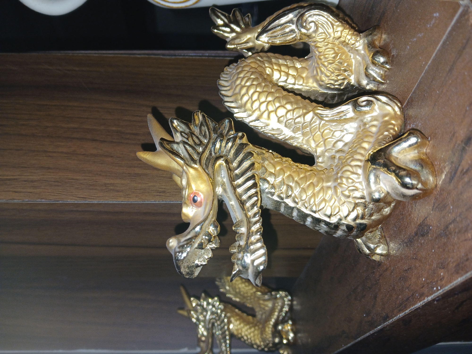 A golden dragon statue with intricate details is the focus of the image. It sits on a dark brown, polished surface that shows its reflection. The dragon is positioned horizontally with its head towards the left. It has a long, serpentine body with scales. Its head features ornate spikes, a pronounced snout, and what appears to be a reddish eye. The statue exhibits a high level of craftsmanship, with textures and contours suggesting a sense of movement. The surrounding environment appears to be indoors, with a blurred dark background. Based on visual evidence alone, there is no information suggesting that this image was taken in Bengaluru, India.