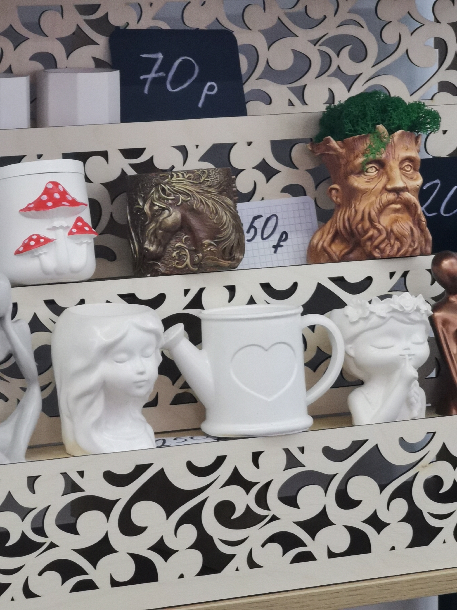 The image shows a shelf in the city of Unknown, Unknown with several decorative items for sale. On the top shelf are geometric white containers, a white container with red mushrooms, a brown container with a horse relief, and a tree-like figure with green moss on top of its head. Small price tags with prices are visible behind the items. The bottom shelf contains a white female figure, a white watering can with a heart, and another white female figure. The shelf has a decorative cut-out pattern.
