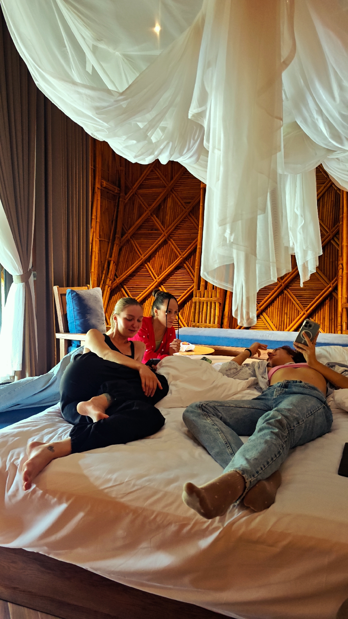 Three young adult females are seen relaxing on a large bed in a bedroom located in Mù Cang Chải, Vietnam. The room features a prominent wall constructed from interwoven bamboo, forming a lattice pattern. A white, sheer canopy hangs over the bed.
On the left, a light-haired female, wearing a black tank top and black pants, is semi-reclined, looking down at a smartphone in her hands. Her bare feet are visible, and a small tattoo is on the top of her left foot. Adjacent to her, in the middle, a dark-haired female in a red top with a distinct collar sits upright, observing something to her right. A small dish is positioned near her. On the far right, a female with medium-length hair is lying on her back, wearing a pink crop top, light blue ripped jeans, and light brown polka-dotted socks. She holds a smartphone with a visible camera array above her head, engaging with the screen. A dark object, possibly another phone, rests on the bed near her feet.
The room is brightly lit, with light entering from a window on the far left, where drapes are pulled back. A wooden chair with a blue cushion is partially visible on the left. The bed is covered with white sheets and pillows.