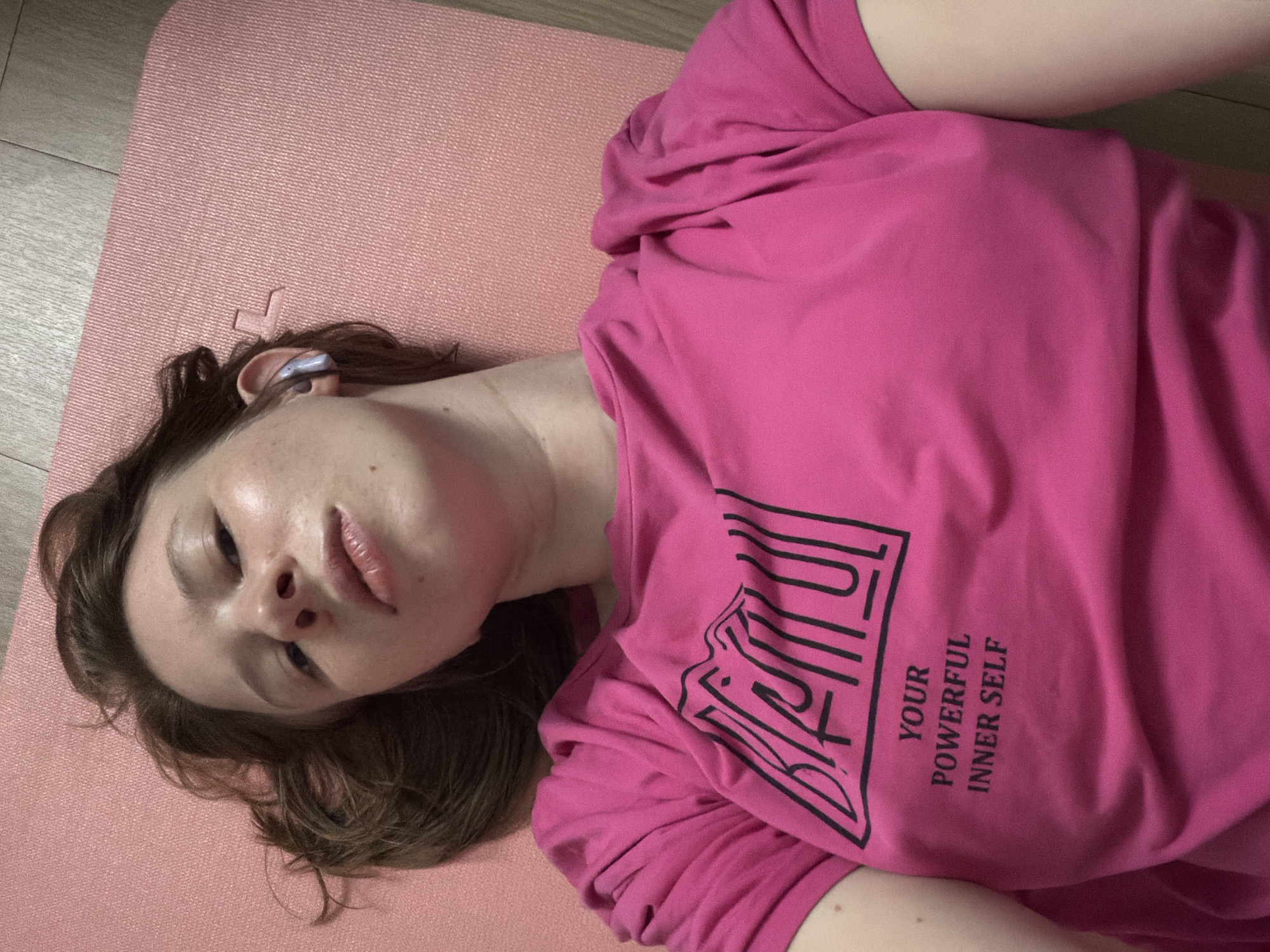 A young woman with short brown hair and fair skin is lying on a pink yoga mat. She is wearing a bright pink t-shirt with black text that reads "BEAUTIFUL" in a stylized font above the words "YOUR POWERFUL INNER SELF." She has earbuds in her ears. The background appears to be a wooden floor and a wall with a pink textured surface. The image seems to have been taken from a high-angle perspective, looking down at the woman. The location is not discernible from the image itself and cannot be determined to be Dongducheon-si, South Korea.