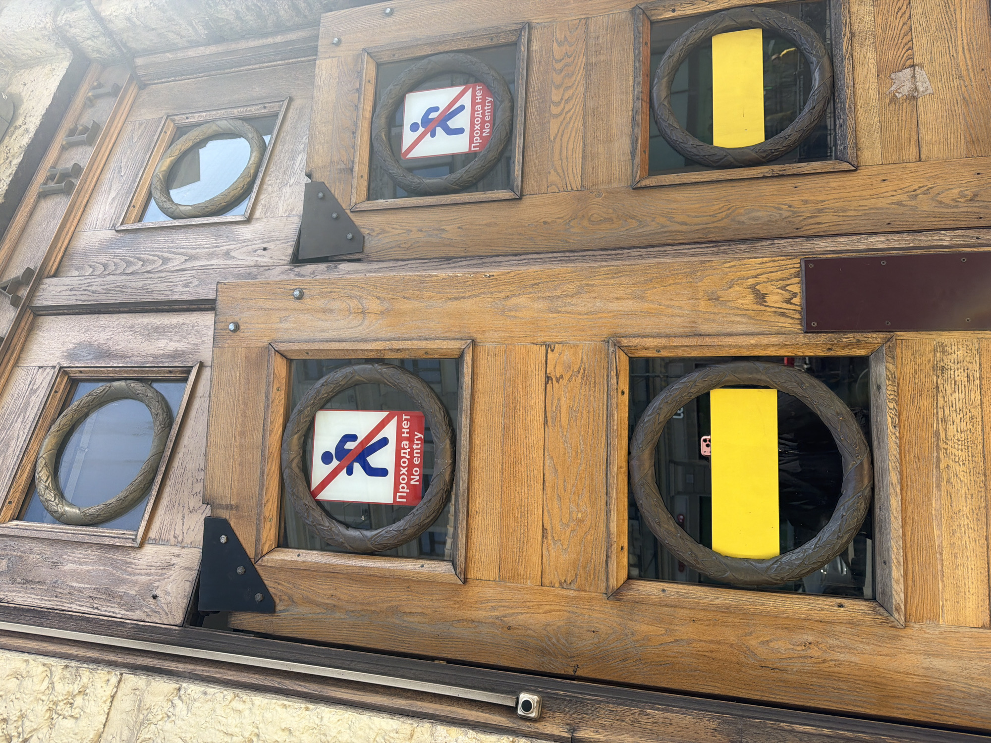 This photograph shows a fragment of a wooden door adorned with circular window frames. Within these frames, various inserts are visible: a clear view through the glass, "No Entry" signs in both Russian ("Прохода нет") and English, and solid yellow inserts. The door's wood grain is prominently visible, and the door hardware includes hinges and screws. The location is Saint Petersburg, Russia.