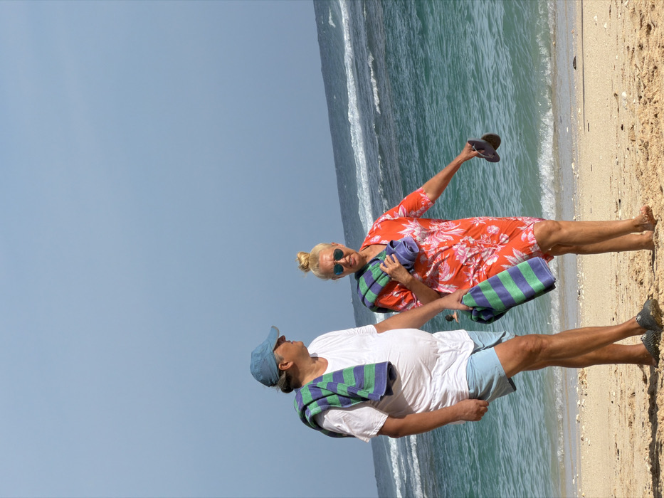 A man and a woman walk side-by-side on a sandy beach in Ungasan, Indonesia, facing left. The man, on the left, wears a light blue baseball cap, a white t-shirt, light blue shorts, and dark slip-on shoes. A green and purple striped towel is draped over his left shoulder. He is looking towards the woman. The woman, on the right, wears a red floral pattern dress, sunglasses, and has her blonde hair in a bun. A similar green and purple striped towel is draped over her left shoulder and arm. She is barefoot and holds a pair of dark flip-flops in her right hand. The background features turquoise ocean water with breaking waves and a clear, light blue sky. A blurred individual is partially visible in the water between the two subjects.