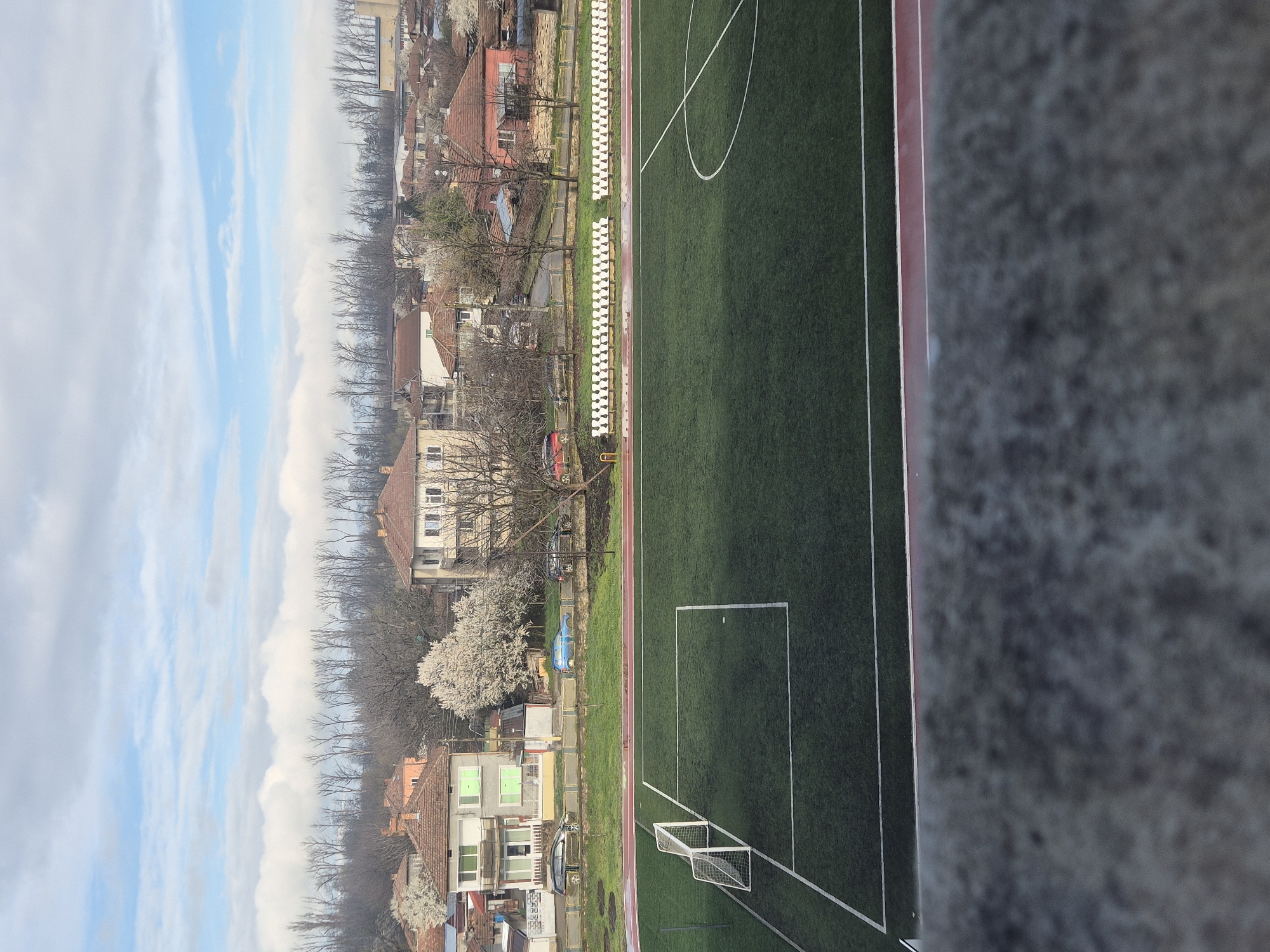 The image shows a soccer field in Pavlikeni, Bulgaria, viewed from an elevated perspective. The field is green with white lines marking boundaries and goal areas. A soccer goal is visible. Adjacent to the field is a row of white seats and a running track. Beyond the track, there are residential buildings with orange roofs and bare trees. The sky is partially cloudy with patches of blue. A concrete structure is visible in the foreground.