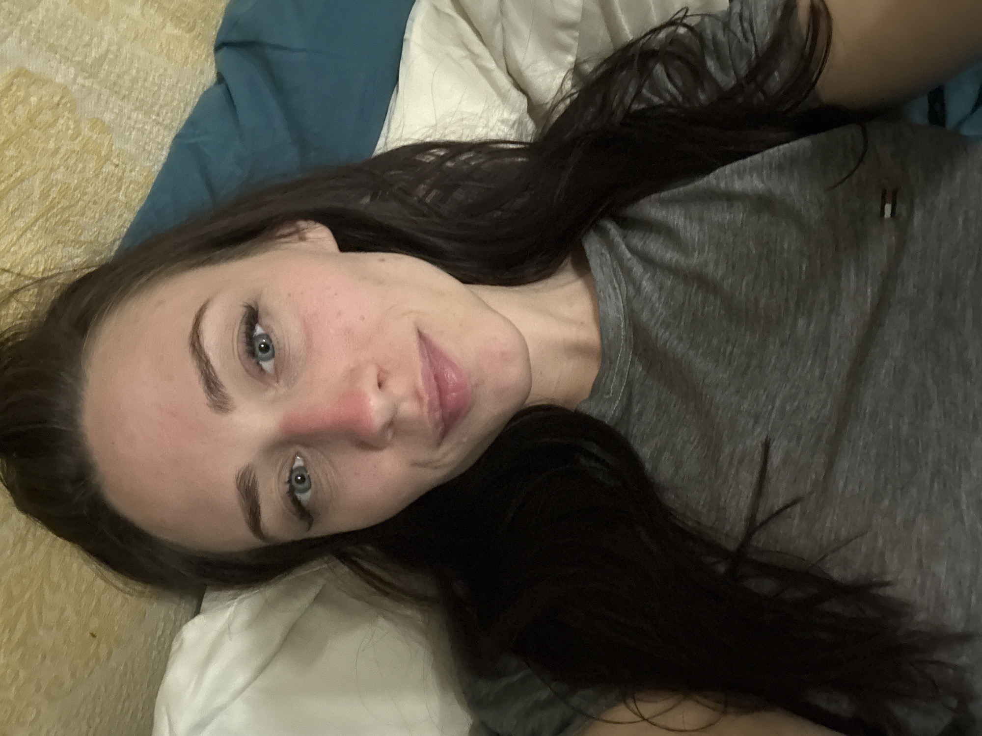 A woman with dark hair and blue eyes is lying on a bed. She is wearing a gray t-shirt with a small red and white logo on the upper right chest area. The bed has white sheets and a blue blanket. The background appears to be a tan wall or a textured fabric. The location is indeterminate, and there is no evidence to suggest the image was taken in Saint Petersburg, Russia.