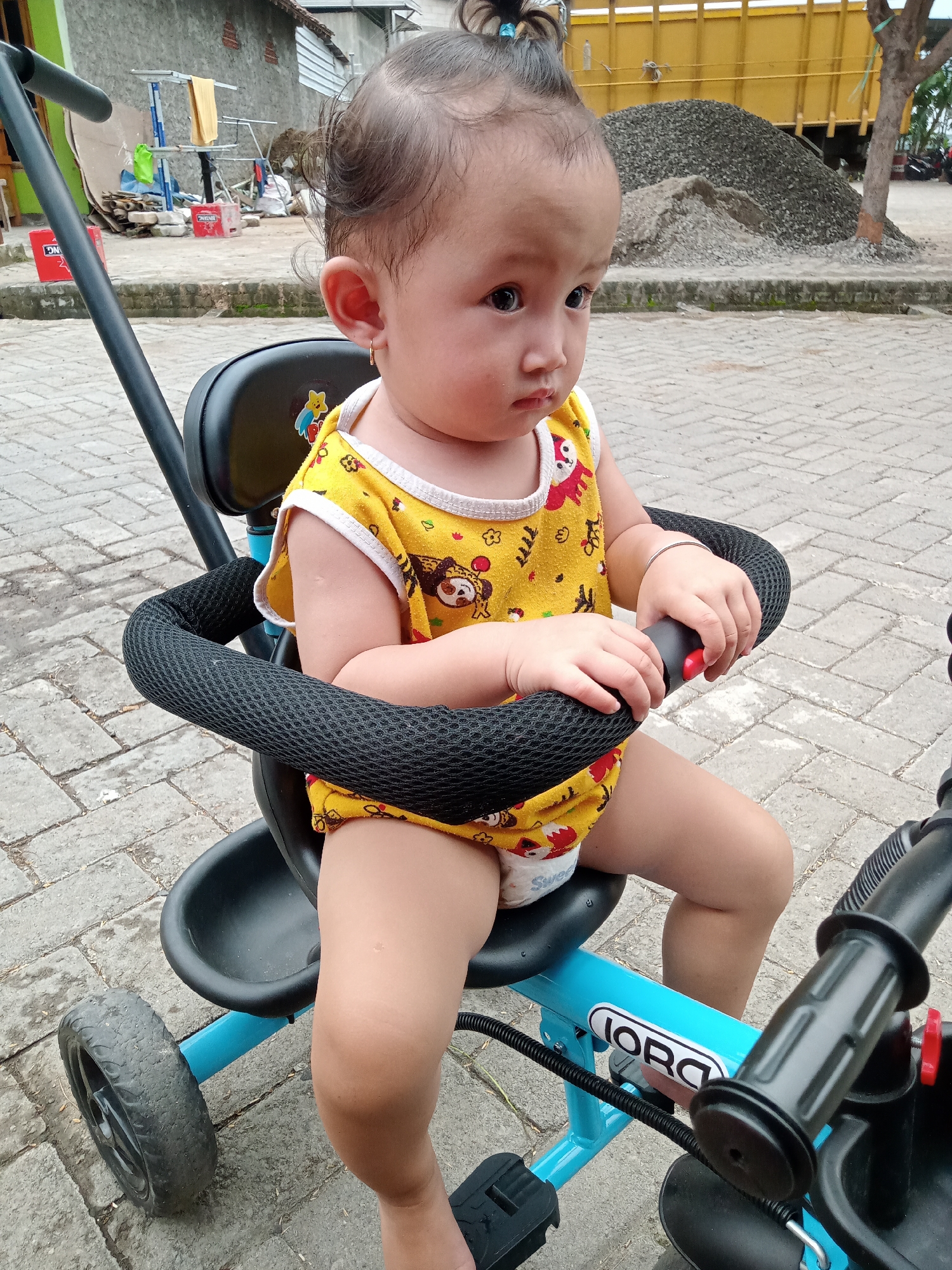 A baby or toddler with dark hair tied in a top bun is seated in a blue and black tricycle-stroller outdoors. The child wears a yellow sleeveless onesie with cartoon animal patterns and white trim, along with a disposable diaper. A small golden hoop earring is visible in the baby's left ear, and a thin silver bracelet is on the right wrist. The baby holds onto the padded black safety bar/handlebars of the tricycle, looking pensively towards the right side of the frame.
The scene is set on a paved surface of grey interlocking bricks. In the background to the left, there's a grey building or wall, a blue laundry drying rack with yellow fabric, and red plastic crates, one bearing the letters "DN". To the right, further back, a large pile of grey gravel dominates, with a yellow truck partially visible behind it, and a tree trunk to its right. The outdoor lighting suggests bright daylight. The location is Bungur, Indonesia.
The word "JORO" is visible in white letters on an oval sticker on the blue frame of the tricycle. The word "SWEET" is partially visible on the baby's diaper. The image captures a quiet moment of observation in what appears to be a residential or semi-urban environment.