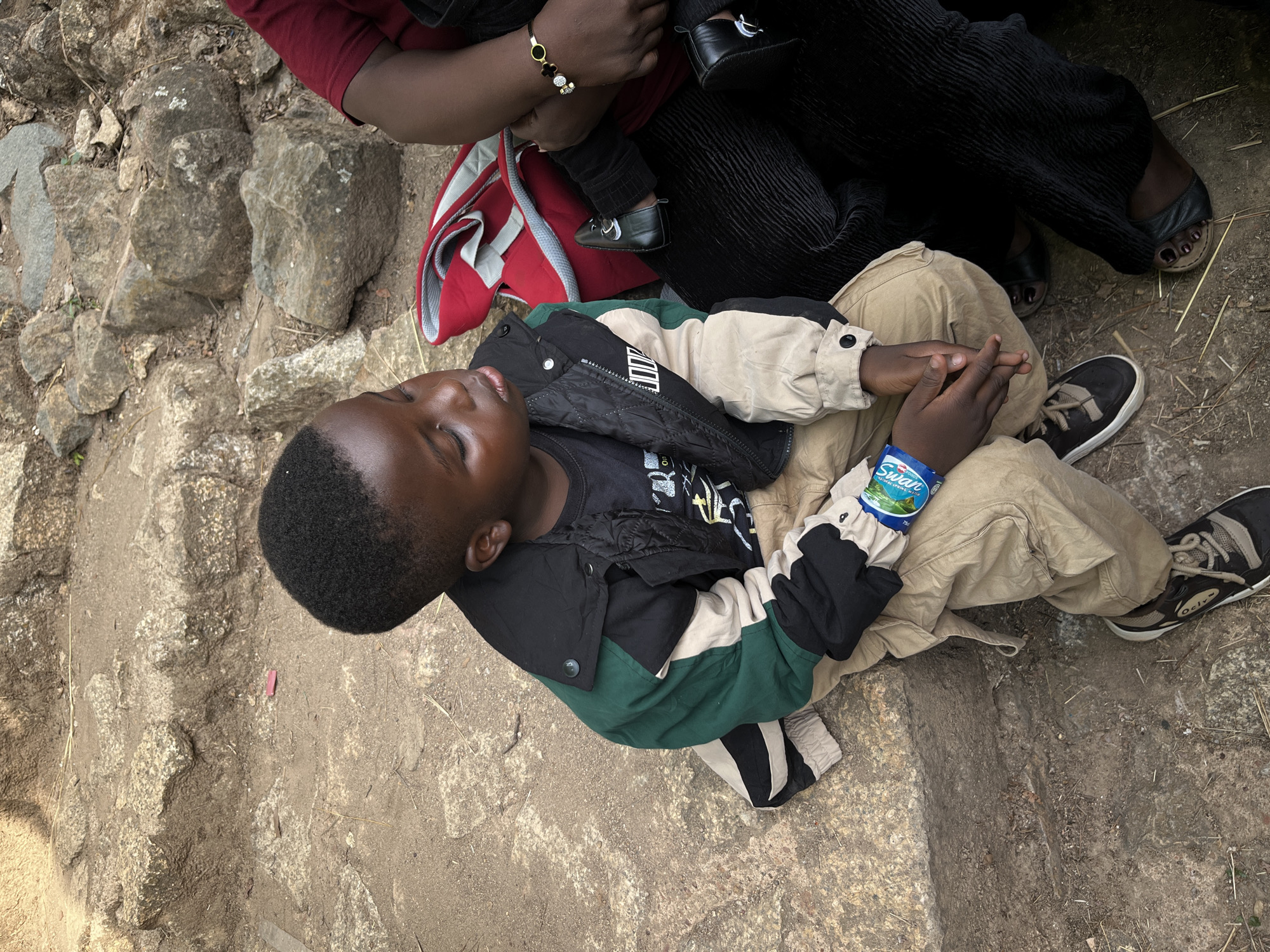 A young Black boy lies on rocky, dusty ground, appearing to rest or sleep with his eyes closed and lips slightly parted. He is dressed in a black t-shirt, a quilted black vest, a green, black, and beige color-block jacket, and khaki pants. His hands are clasped over his stomach area, and a small blue and white packet labeled "Swan" is tucked under his right wrist. He wears black and beige sneakers with white soles, and the side of one shoe shows text, possibly "Oclyx" or "Oclix."
Above the boy, an adult's legs and feet are visible, clad in black ribbed pants and black sandals, with dark-painted toenails. This adult appears to be sitting or crouching. Adjacent to them, a red and grey backpack is on the ground. A hand and arm, possibly belonging to the same adult, are seen holding a baby or toddler whose legs are dressed in black pants and black shoes with white detailing. A gold bracelet with charms is on the adult's wrist.
The setting is outdoors, characterized by uneven, dry, and rocky terrain with scattered small stones and patches of sparse dry vegetation. The lighting is soft and diffused, suggesting either an overcast day or shade. A small pink object is on the ground near the boy's head. The scene depicts a moment of rest or waiting for a small group, likely in Jos, Nigeria.